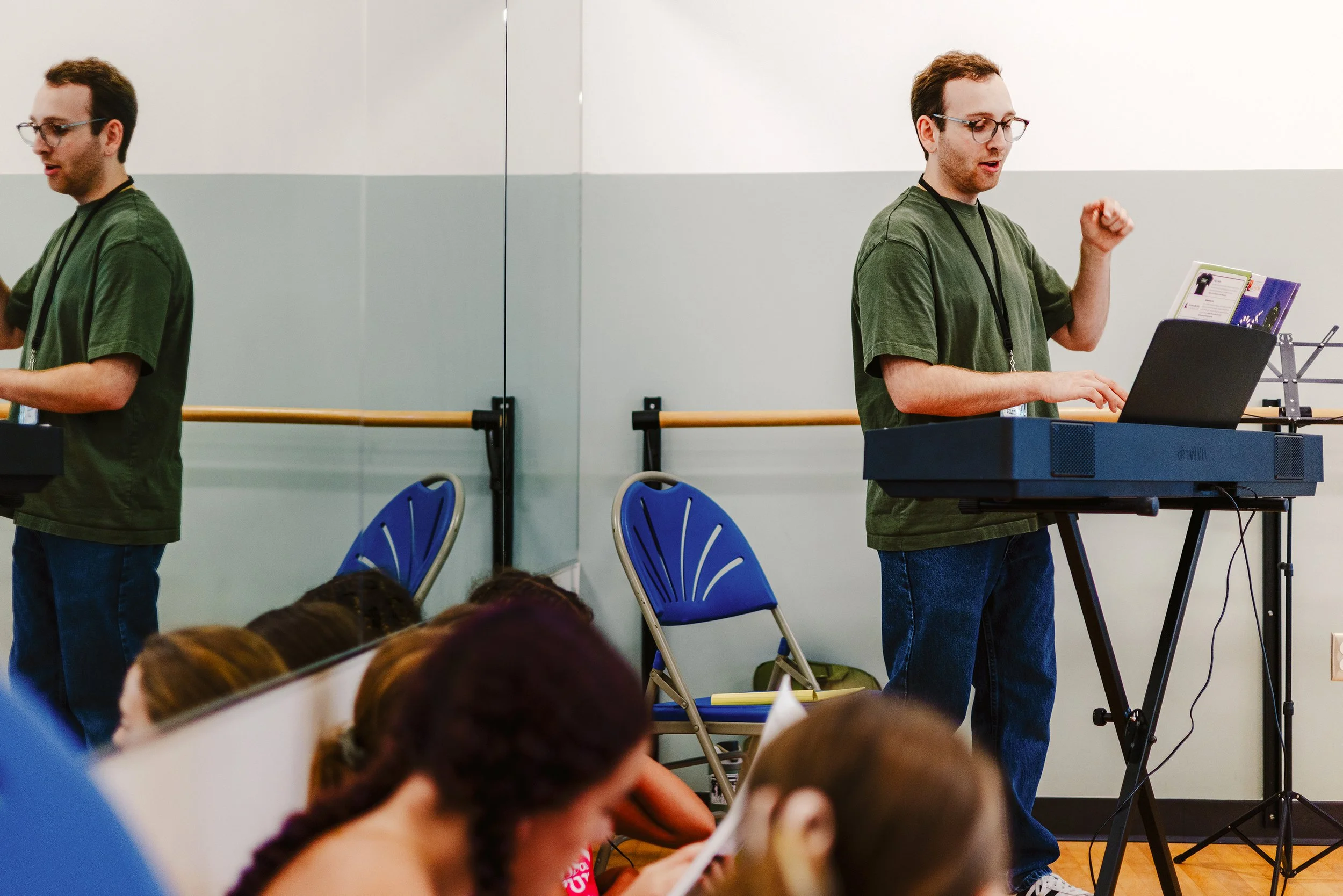 A man in a green shirt playing a digital keyboard with a laptop in front of him, while a man on the left side of the image appears to be playing an instrument or reading music, with people sitting in the foreground. The setting looks like a classroom or a rehearsal space with light-colored walls and chairs.
