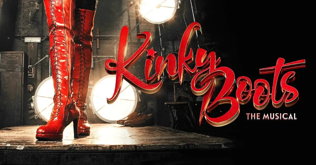Red knee-high boots with laces on a stage with a spotlight and the sign 'Kinky Boots The Musical'.