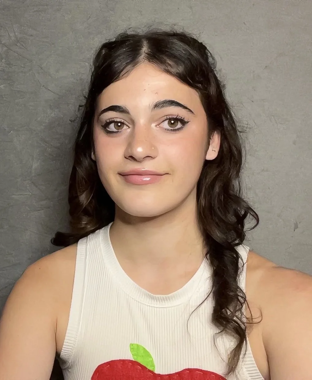 A young woman with dark brown, curly hair and light skin taking a selfie against a gray wall. She has makeup on, including eyeliner and glossy lips, and is wearing a sleeveless cream top with a red apple graphic.