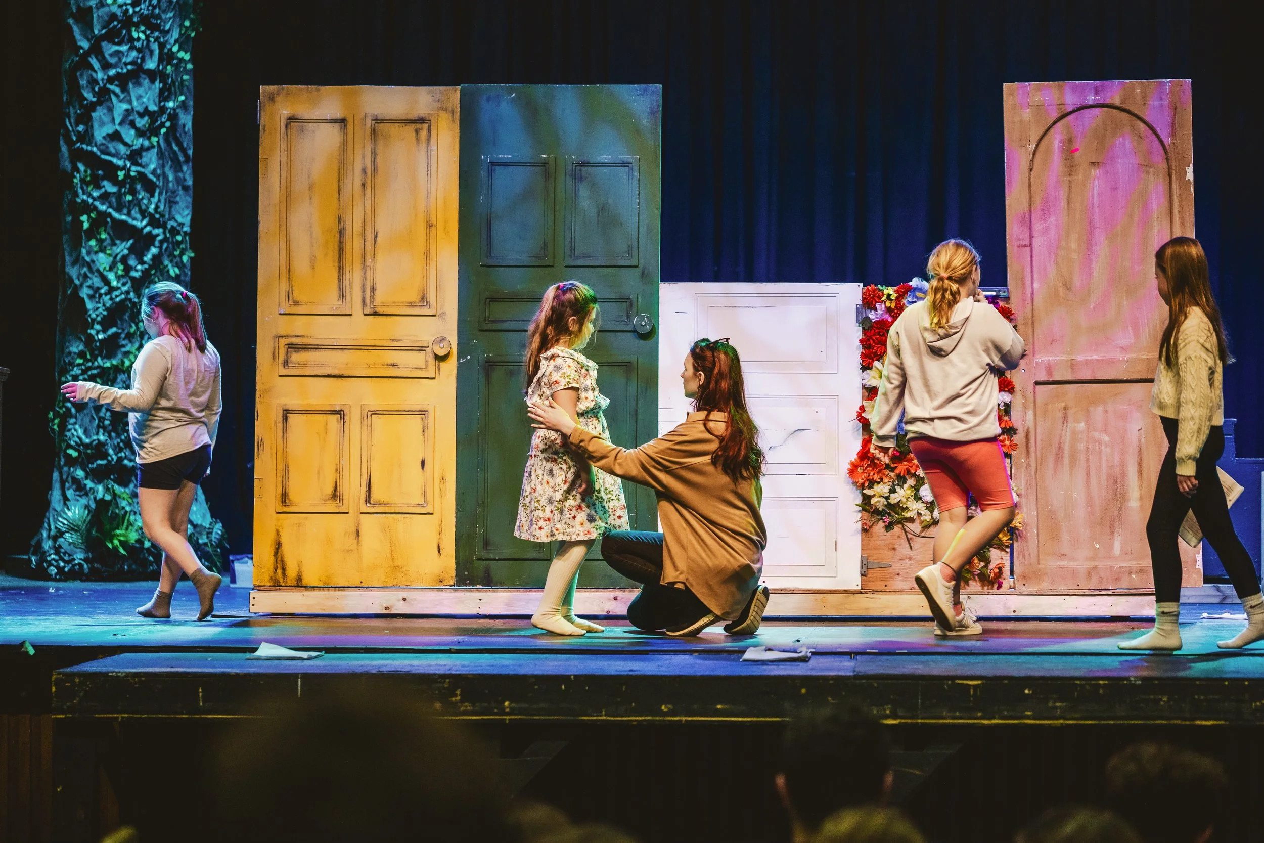 Young girls rehearsing a play on stage with colorful doors and floral decorations, one girl kneeling and talking to another, others walking.