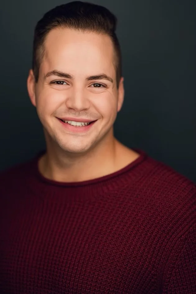 Portrait of a young man with short dark hair, smiling, wearing a maroon sweater, against a dark background.