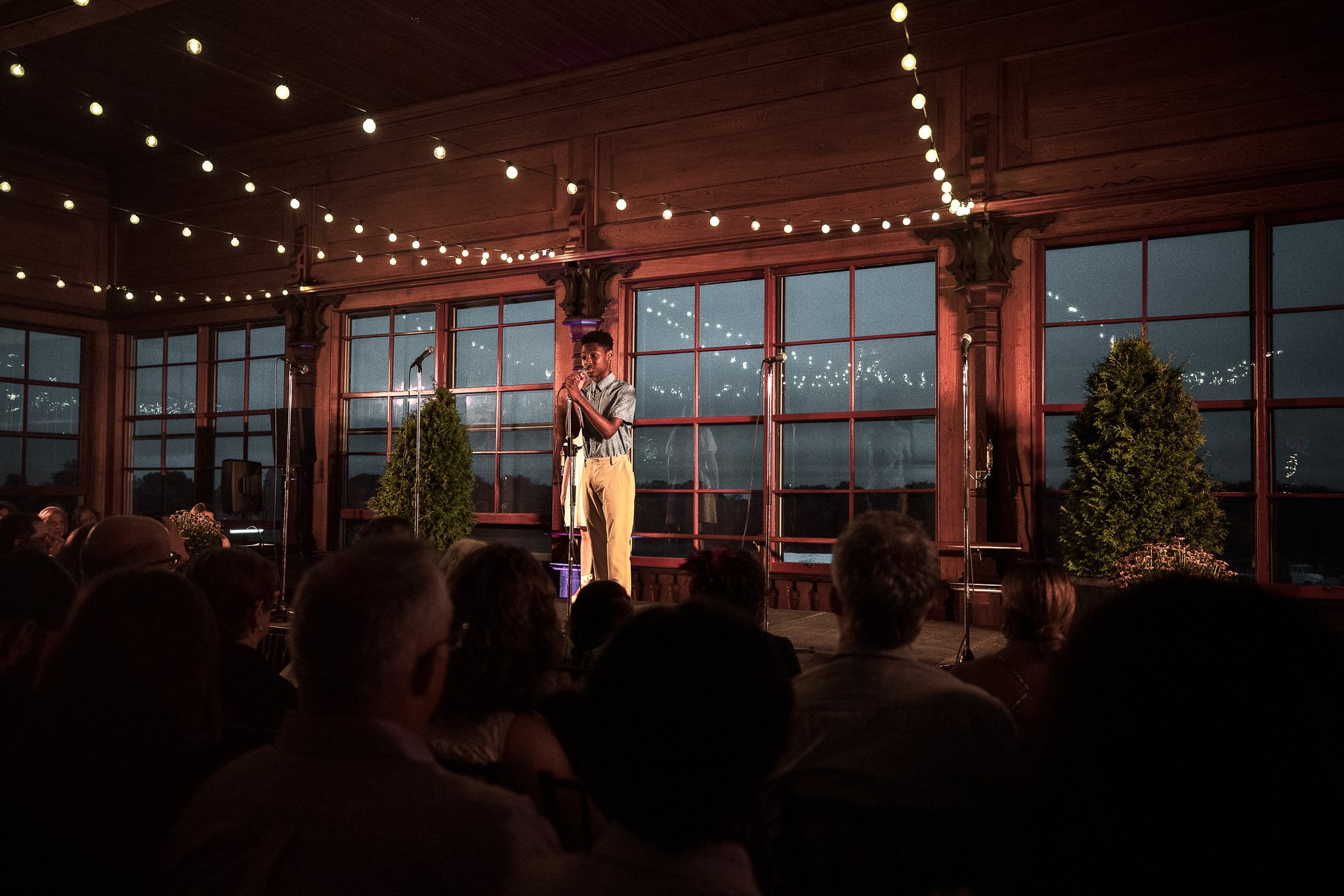 A person standing on a stage, speaking or performing into a microphone, with an audience seated in front. The stage has large windows, potted plants, and string lights hanging from the ceiling, creating a cozy, indoor event setting during the evening or night.