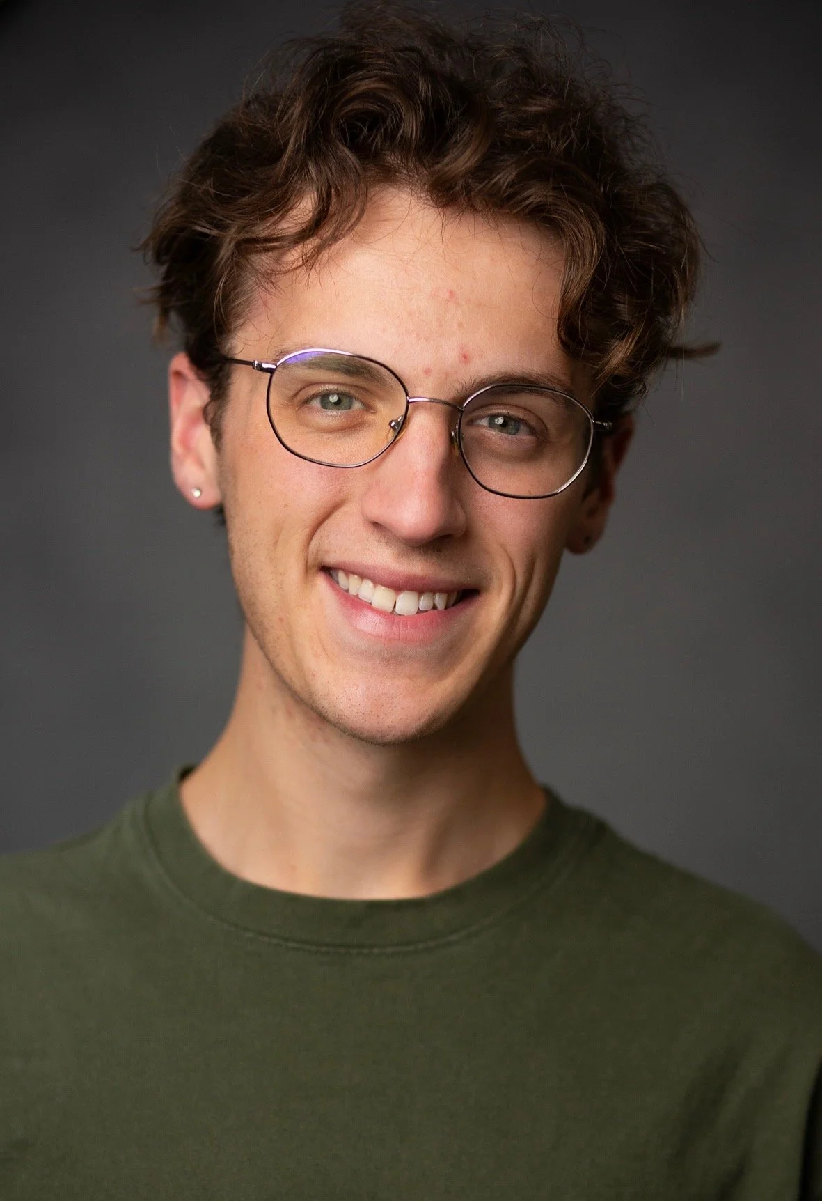 A young man with brown hair, glasses, and earrings, wearing a light-colored, short-sleeved, patterned shirt, standing in front of green foliage.
