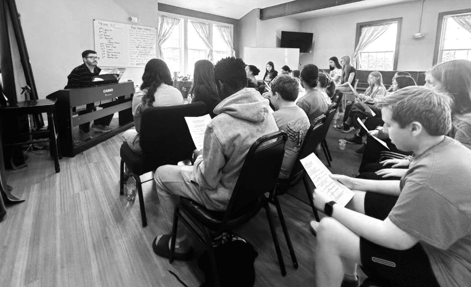 A group of students attending a music class or rehearsal in a classroom with a teacher at a keyboard, with students seated and holding papers, some reading, in a bright room with large windows and curtains.