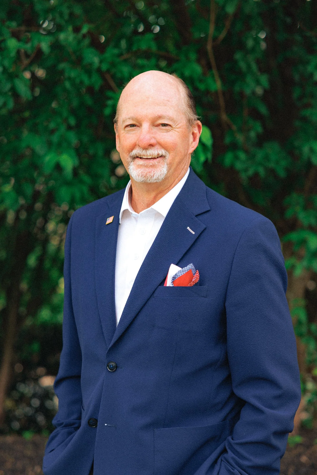 A smiling man in a blue suit with an American flag pin, white shirt, and red pocket square, standing outdoors with greenery in the background.