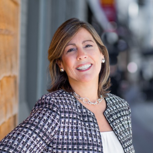 Professional woman outdoors smiling, wearing a patterned blazer, pearl earrings, and a silver necklace.