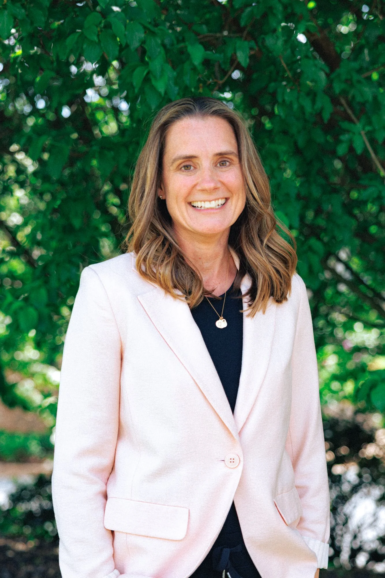 A woman with long brown hair, smiling, wearing a white blazer over a black top, standing in front of green foliage.