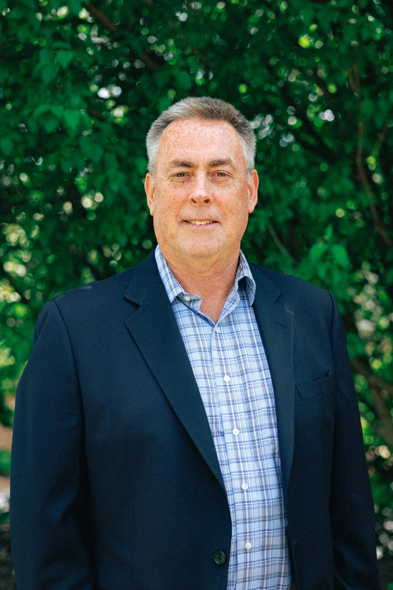 A middle-aged man with short gray hair, wearing a dark blazer over a blue plaid shirt, standing outdoors in front of green foliage.