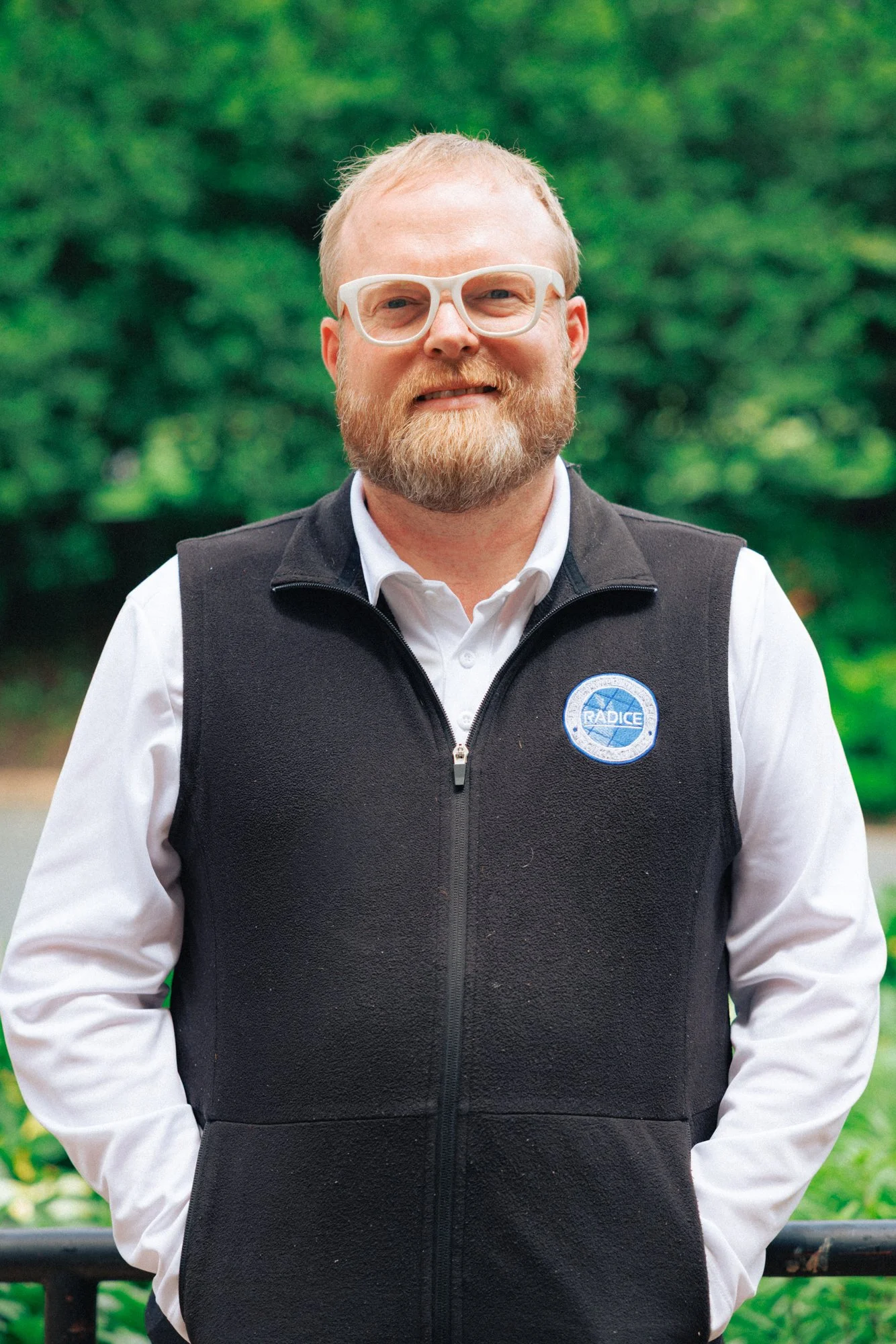 A man with short blond hair, glasses, and a beard, wearing a black vest with a logo, standing outdoors with green trees in the background.