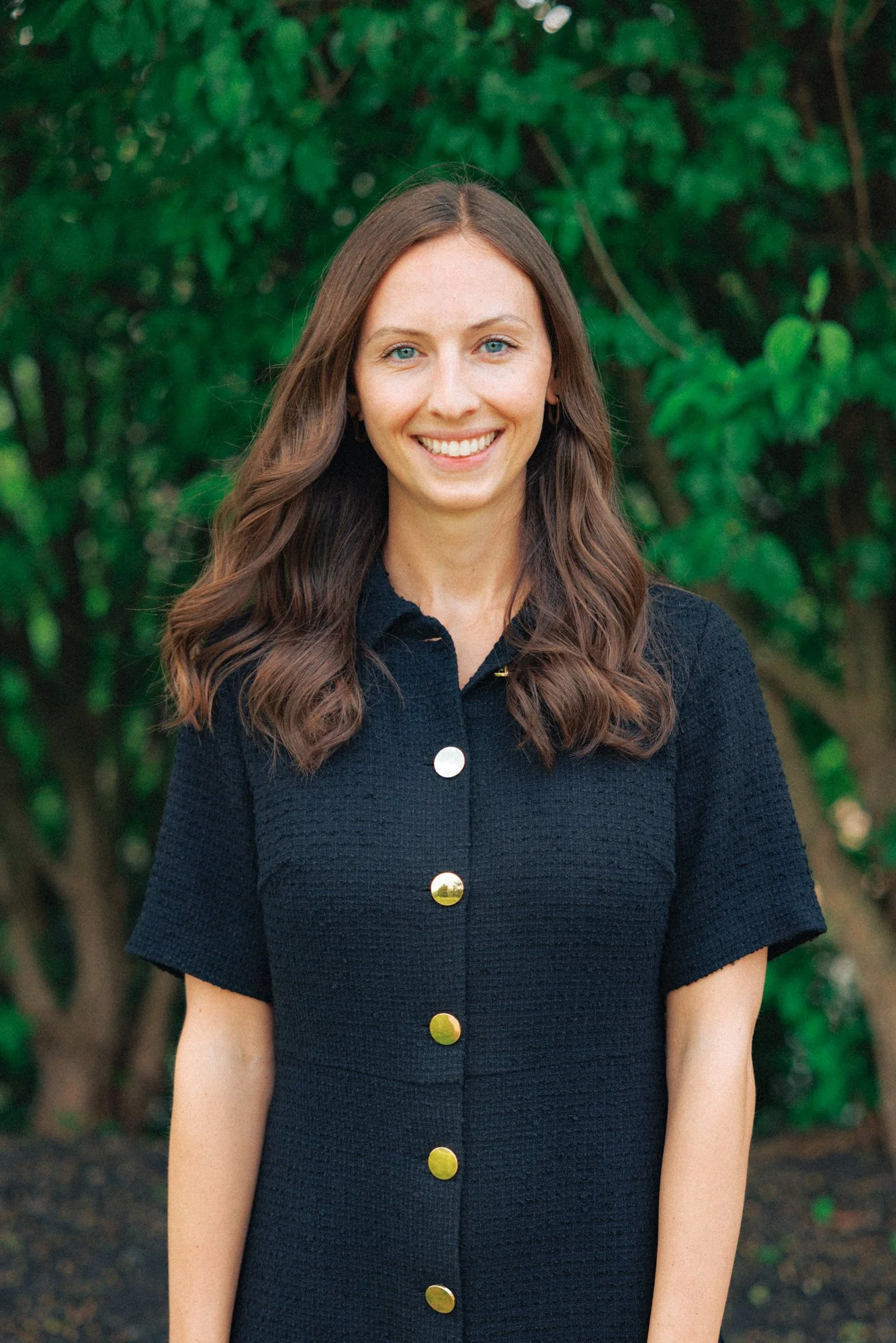 A woman with wavy brown hair and blue eyes, smiling, standing outdoors in front of green foliage, wearing a black textured dress with gold buttons.