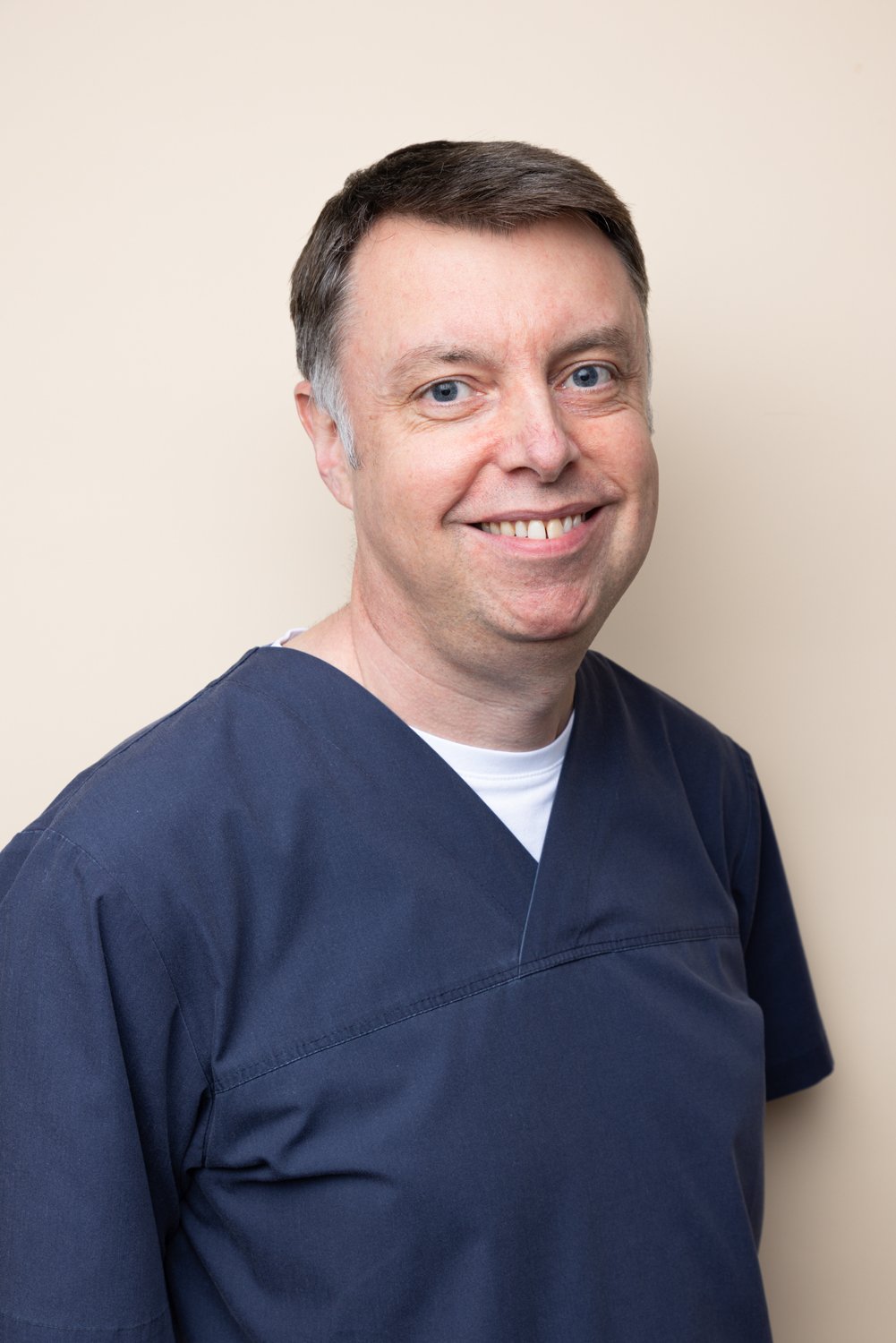 A smiling man with short brown hair, blue eyes, wearing navy scrubs, standing against a plain light-colored background.