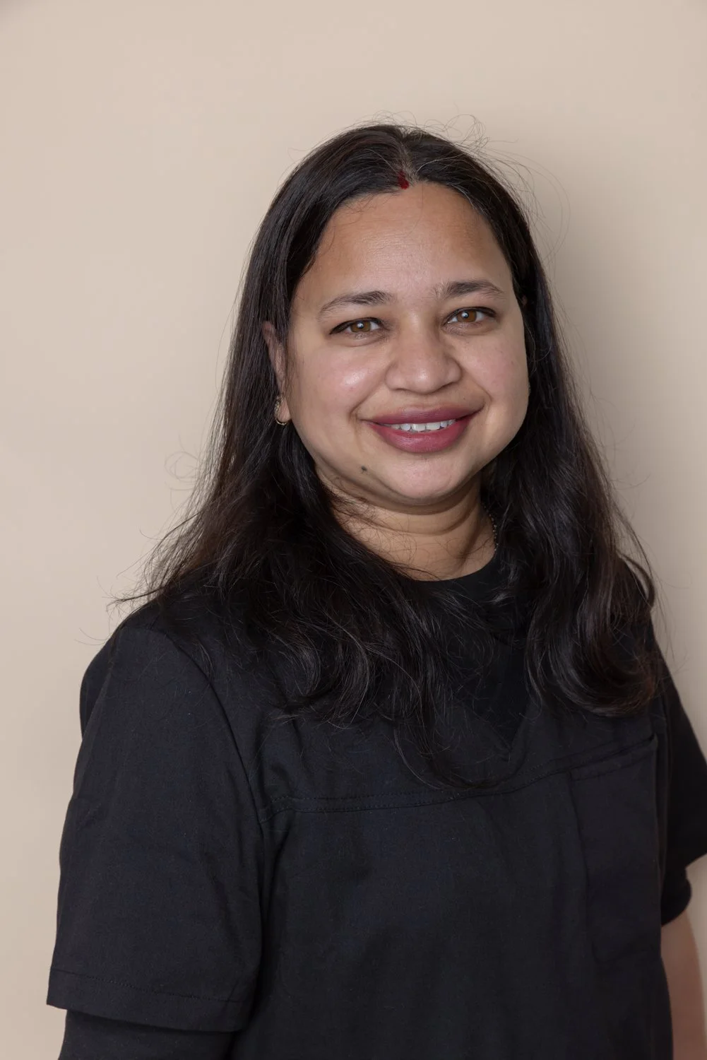 A woman with long dark hair, wearing a black top, smiling, standing against a plain beige wall.
