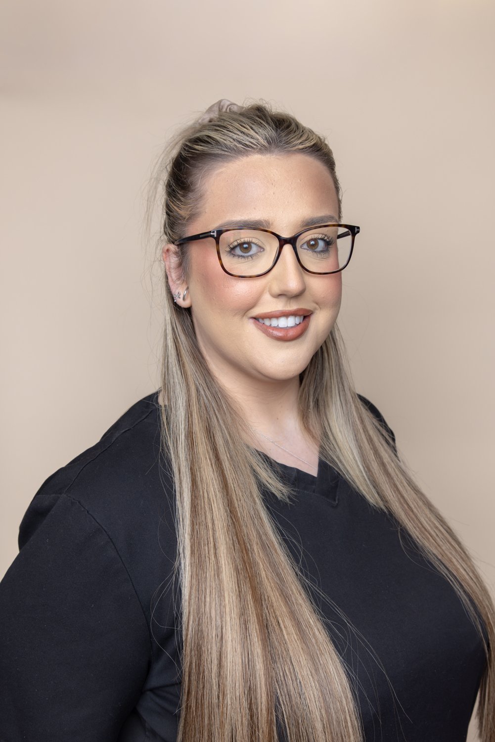 A woman with long blonde hair, wearing glasses and black scrubs, smiling against a beige background.