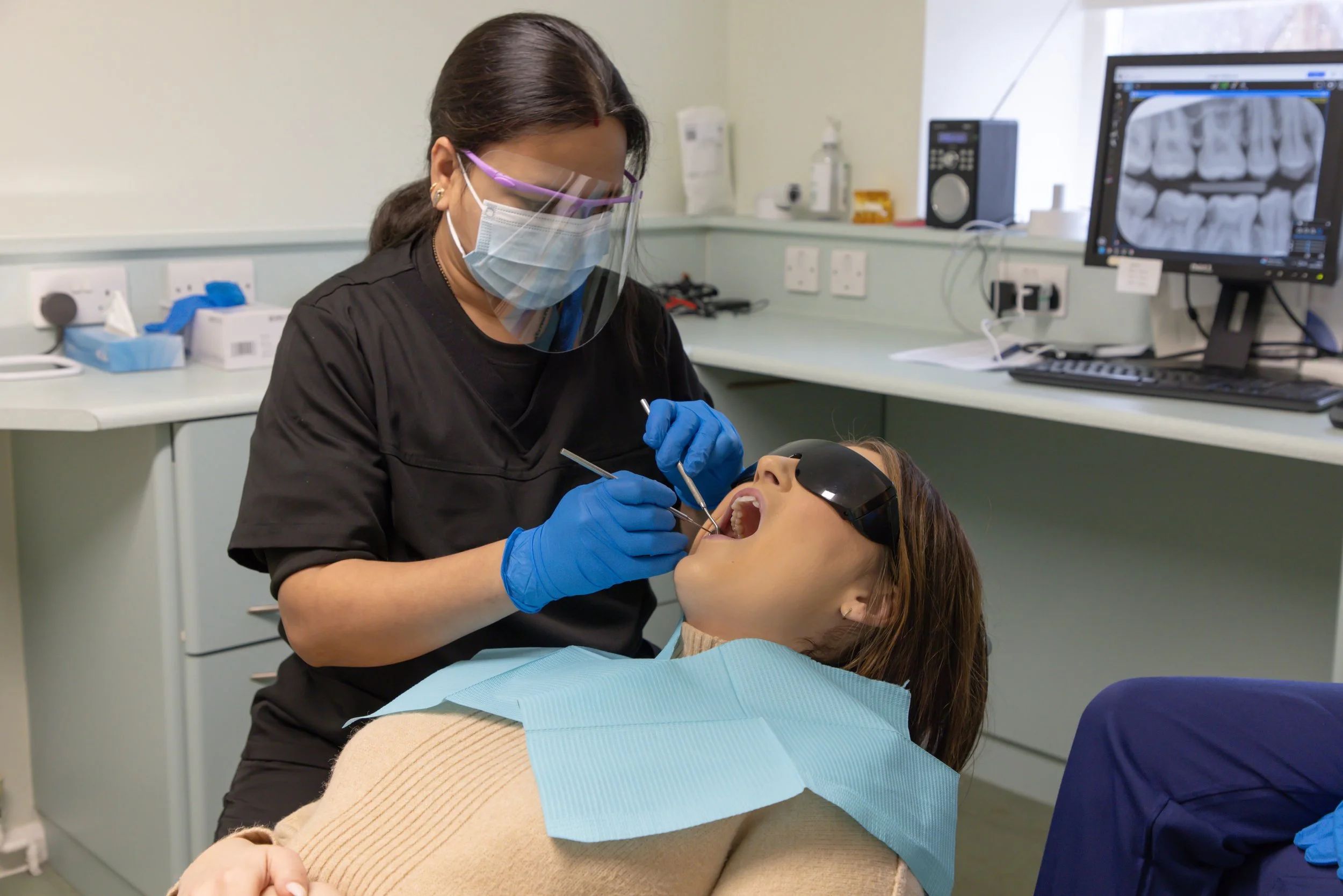 A dentist wearing a face shield, mask, and gloves is examining a patient's mouth in a dental clinic, with dental x-ray images displayed on a computer screen in the background.