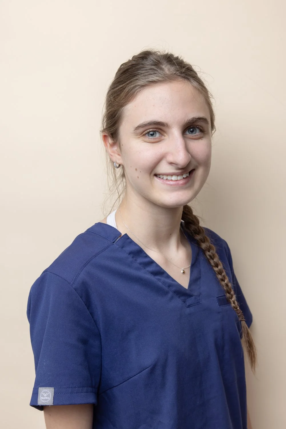 Portrait of a young woman wearing a navy blue medical scrub top, smiling and standing against a plain beige wall.