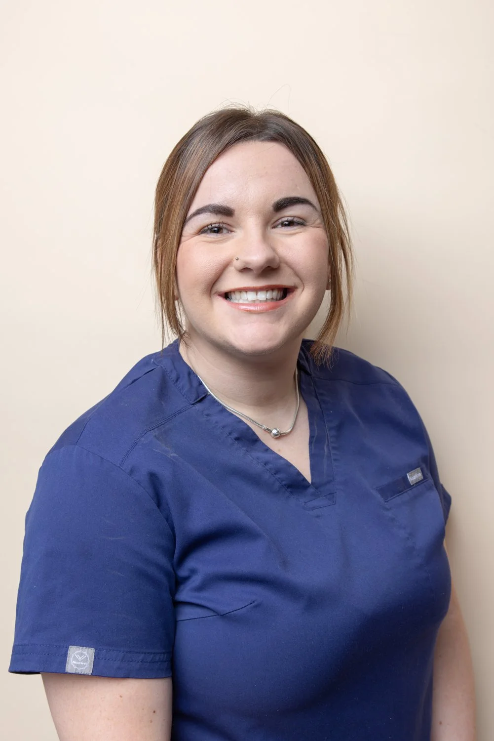 A young woman wearing a blue medical scrub top, smiling and standing against a plain beige wall.