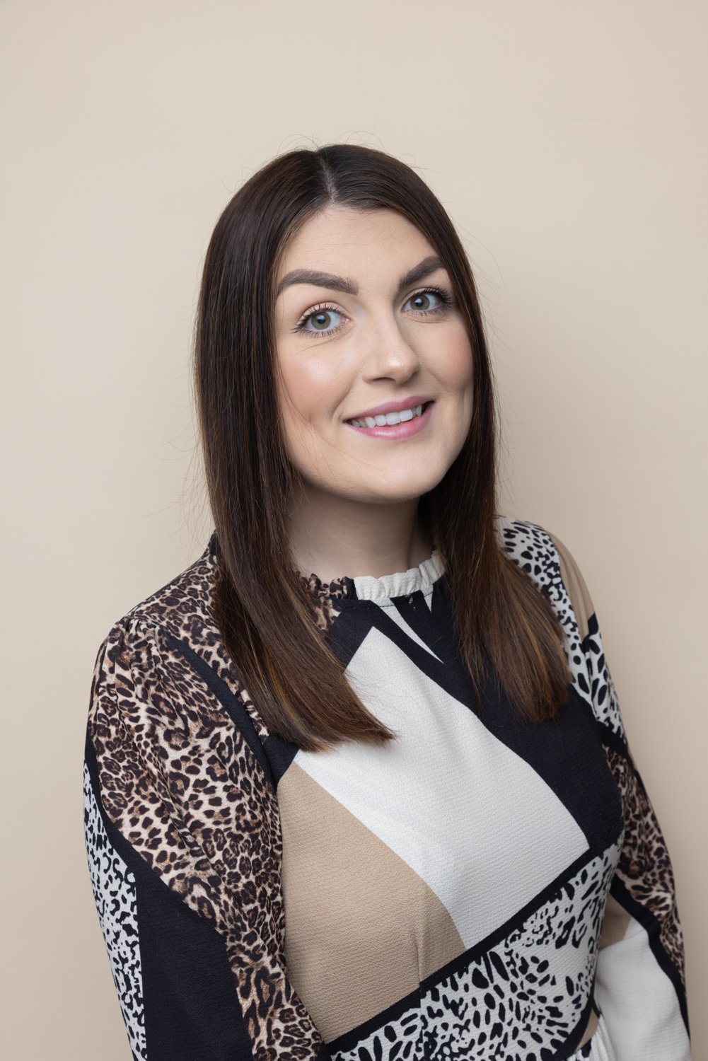 A young woman with shoulder-length brown hair, blue eyes, and a light complexion smiling in front of a beige background. She is wearing a multicolored animal print and geometric patterned top.