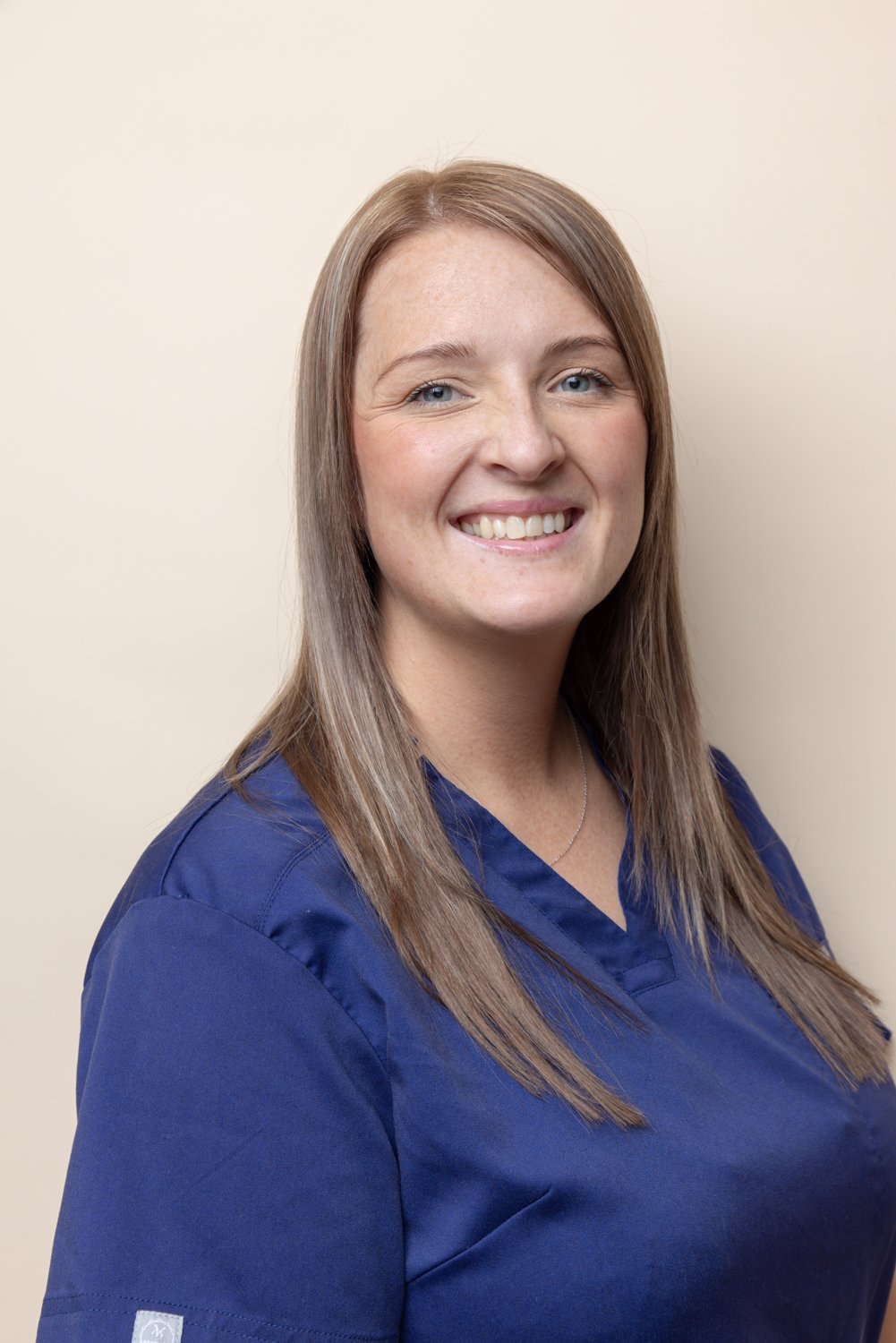 A smiling woman with long, straight, light brown hair and blue eyes, wearing a navy blue scrub top, standing against a beige wall.