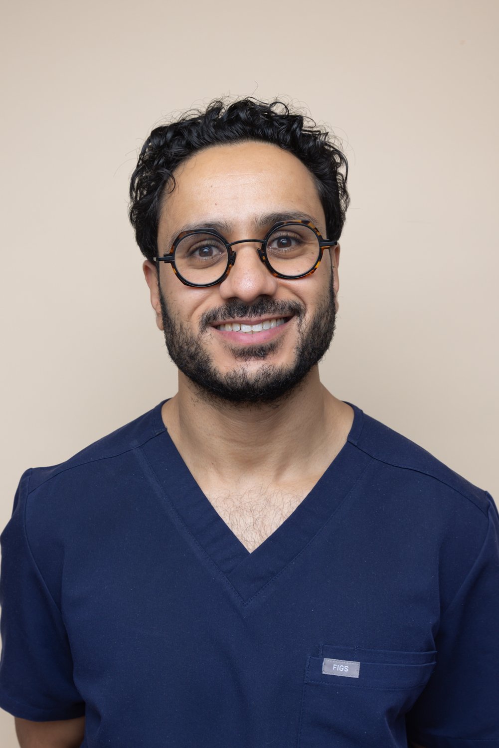 A man with curly black hair, glasses with round tortoiseshell frames, a beard and mustache, smiling, wearing a navy blue scrub top, standing against a plain beige background.