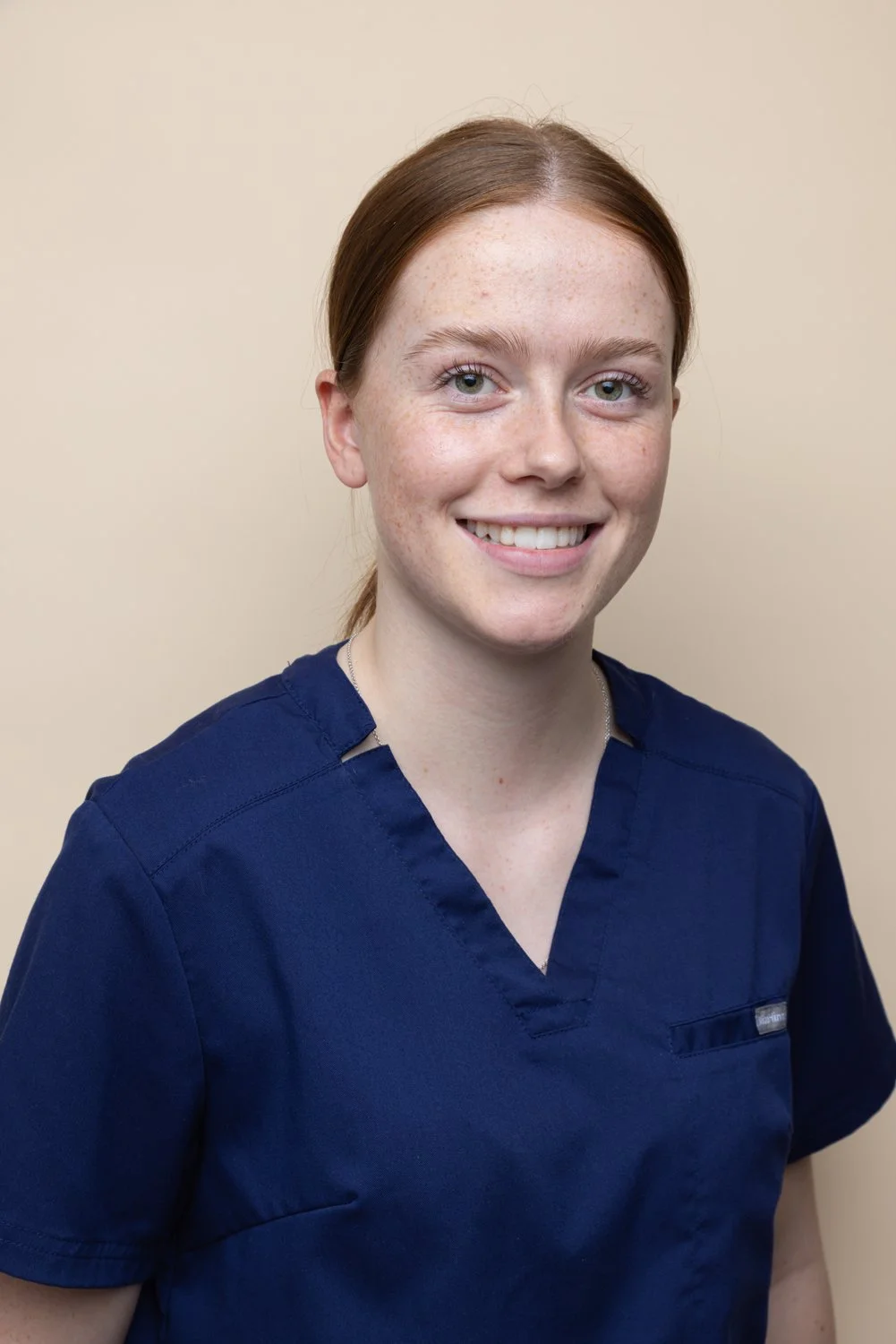 A young woman with red hair, blue eyes, and freckles smiling, wearing navy scrubs against a beige background.