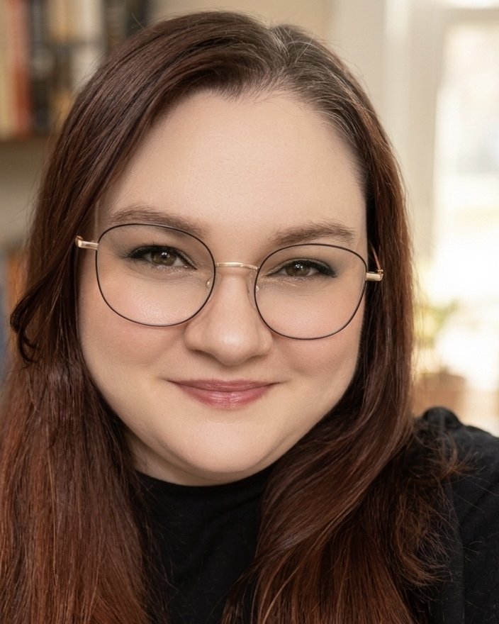 A woman with long wavy reddish-brown hair, wearing glasses with black frames, red lipstick, a black choker, and a black top with a colorful floral pattern on the sleeve. She is smiling softly and is indoors with a light-colored curtain and wooden furniture in the background.