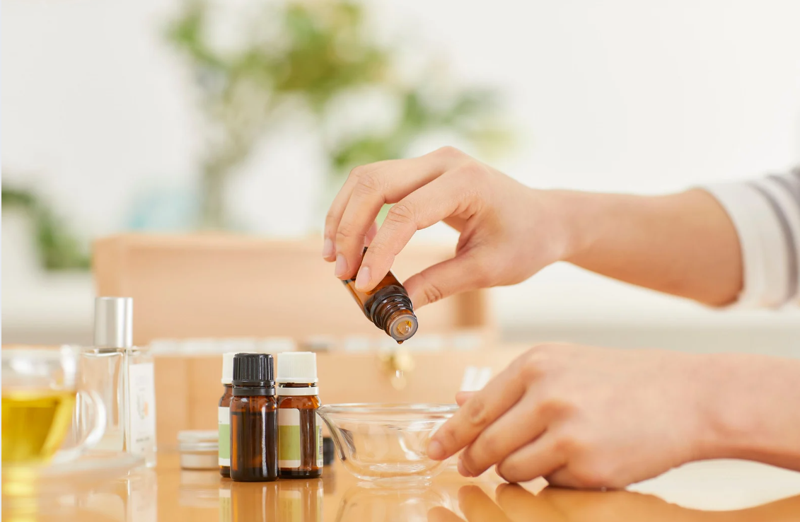 Person pouring essential oil from a small bottle into a glass bowl on a table with other bottles and containers.