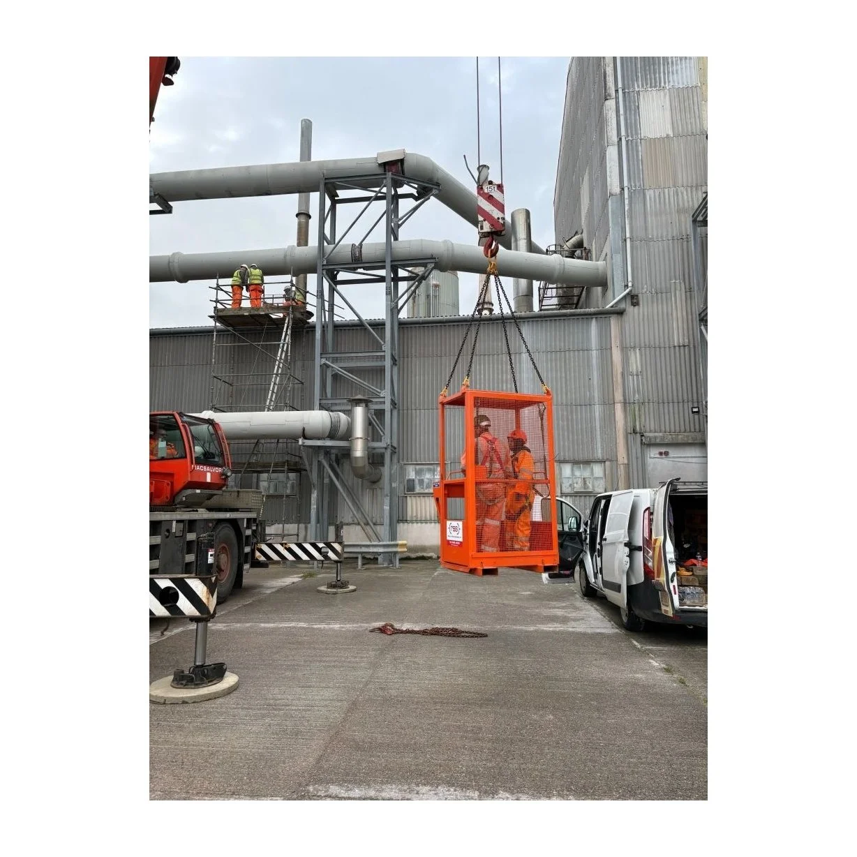 Crane work platform being hoisted by a crane with two people in it. The site is an industrial site. The scale is 1:1