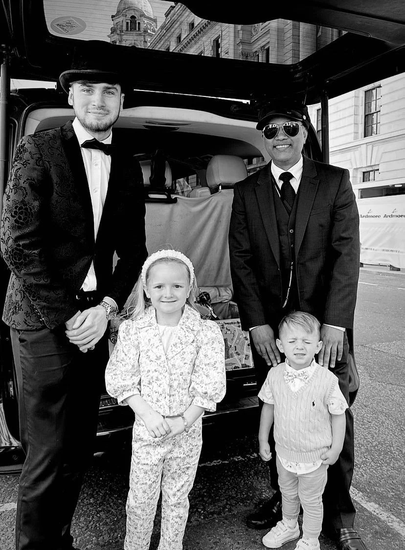 Two men and two children standing in front of a vehicle. The men are dressed in suits, with one wearing sunglasses and a hat. The children are a girl and a boy, with the girl wearing a floral-patterned outfit and the boy in a sweater vest and bow tie. The setting appears to be on city streets with buildings in the background.