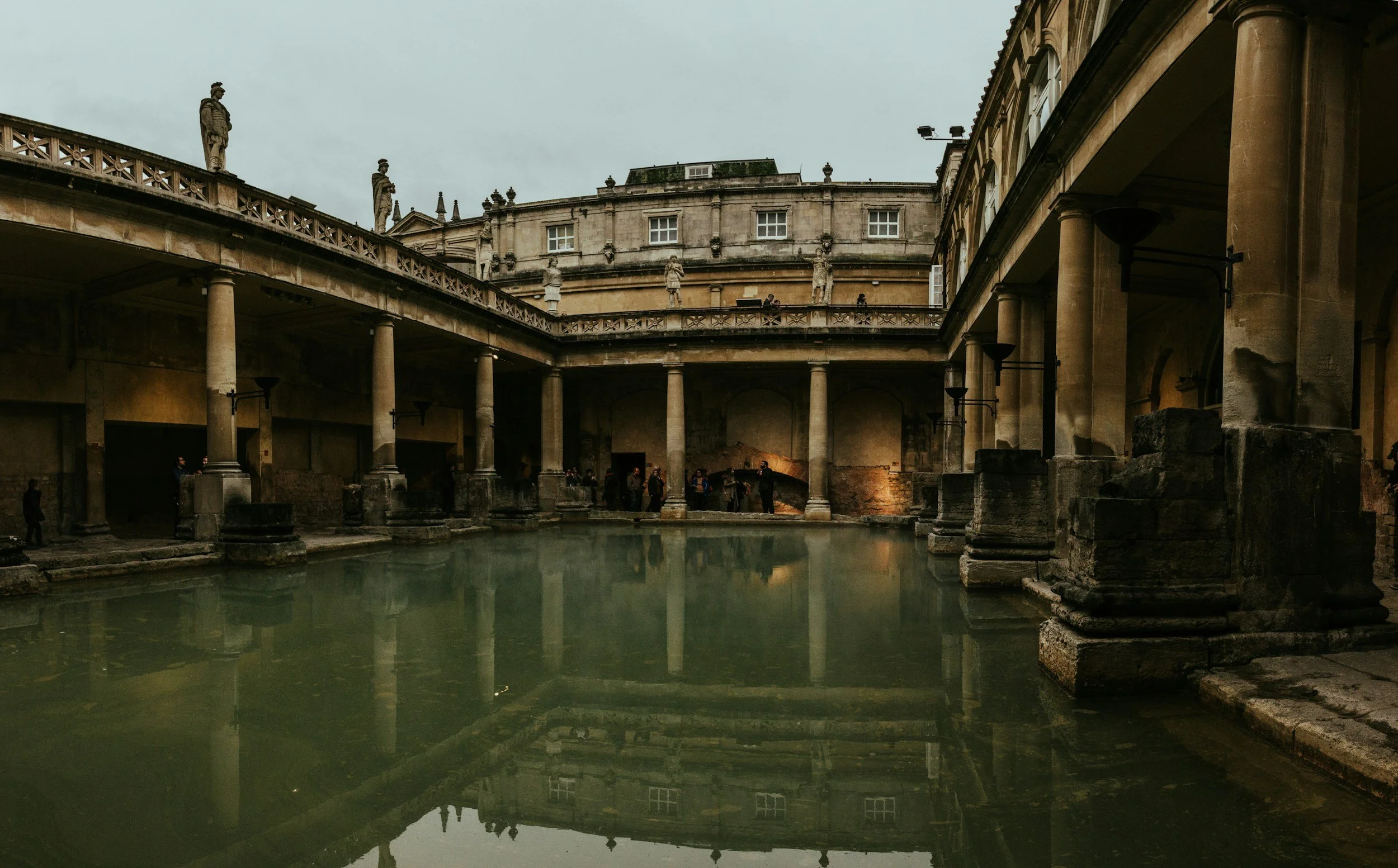 Ancient Roman archaeological site with a rectangular pool of water, surrounded by columns, statues on the upper level, and historic stone buildings under an overcast sky.