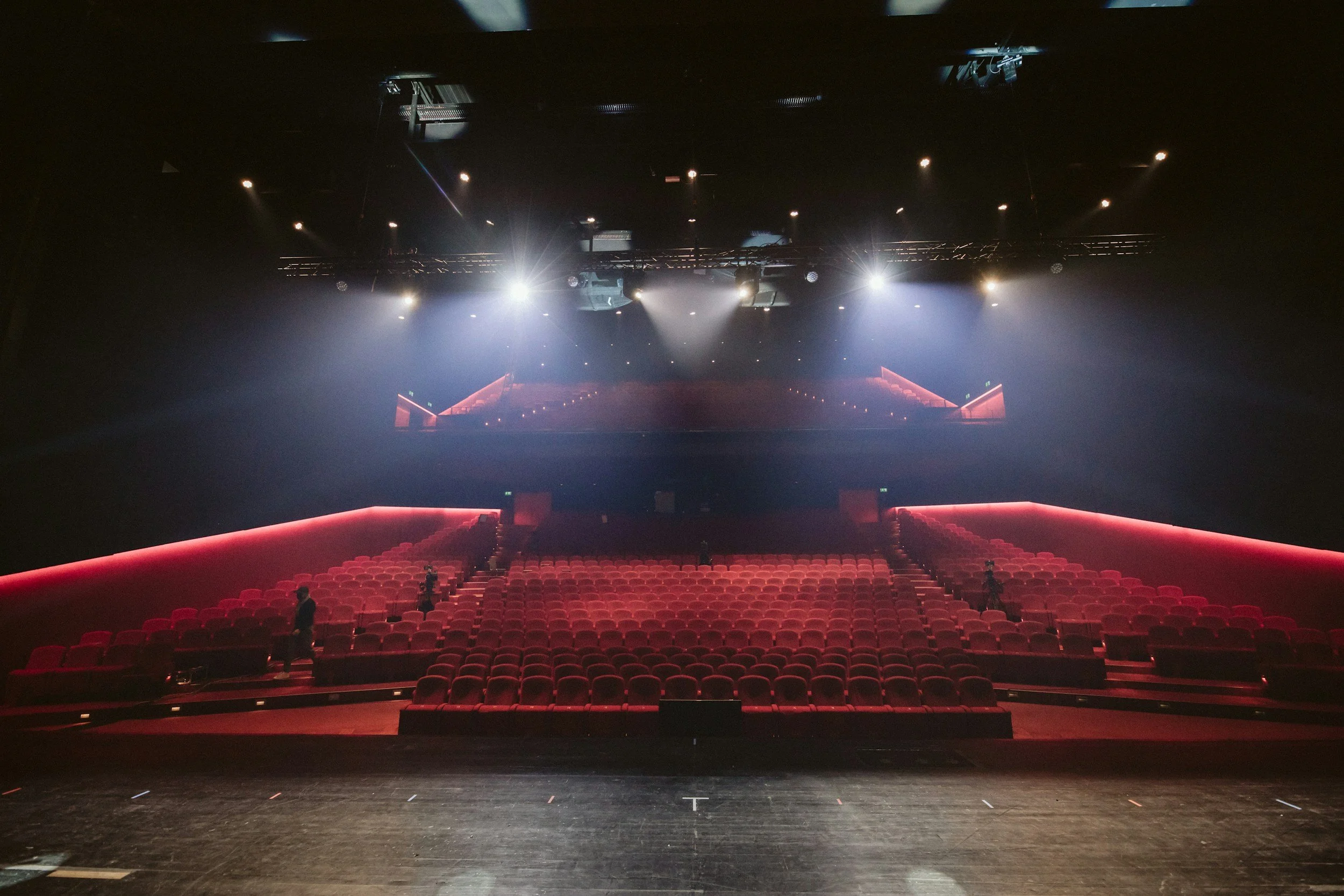 Empty theater with red seats, dim lighting, and spotlights overhead, viewed from the stage.