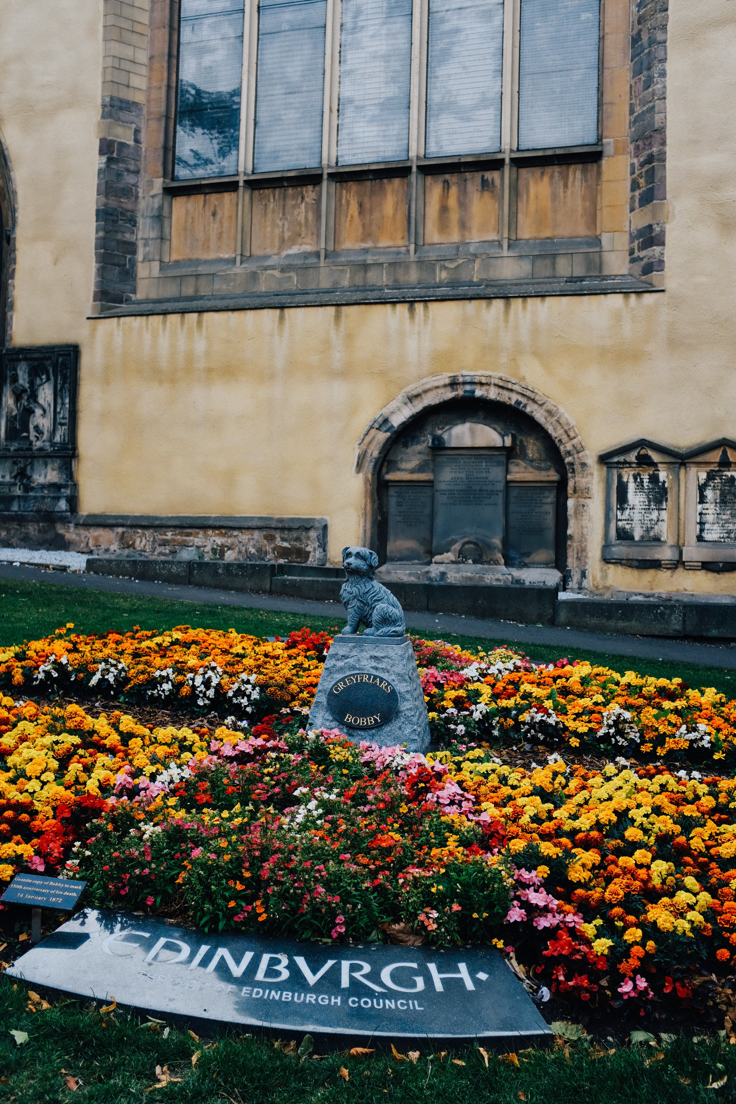 A flower bed with colorful flowers, a small statue of a dog named Bobby, and a plaque with the words 'Gryffyrar' and 'Bobby.' In the background, there is an old stone building with large windows and plaques.