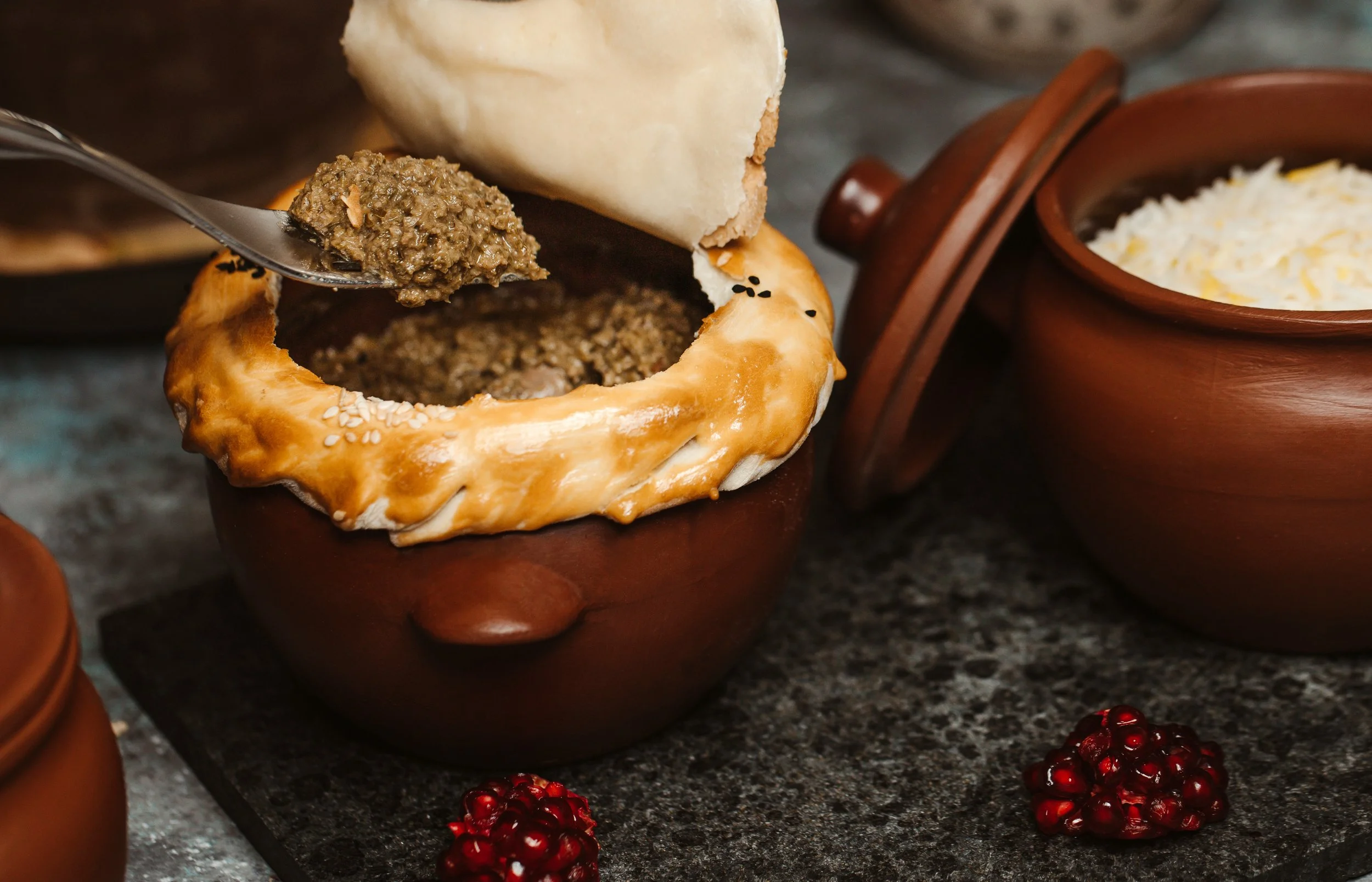 A traditional dish of Ethiopian mesir wat served in a brown clay pot with a bread on top, beside a bowl of shredded cheese, and a small pile of red berries on a dark countertop.