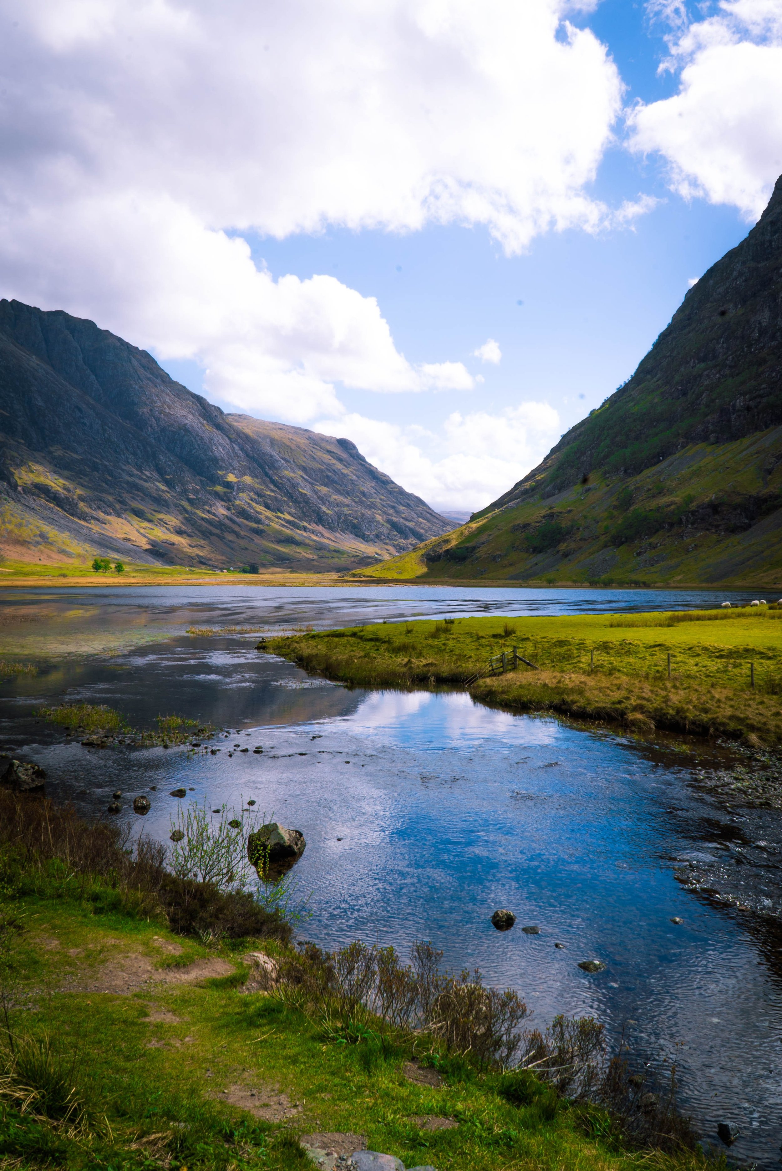 A scenic view of a river flowing through a green valley surrounded by tall mountains with a partly cloudy sky overhead.