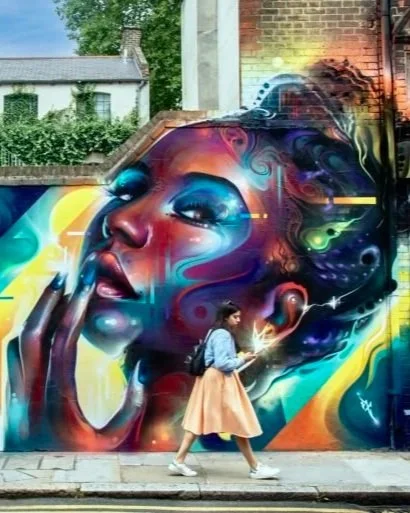 Colorful mural of a woman with vibrant abstract hair, with a young woman walking past holding a phone in front of her.