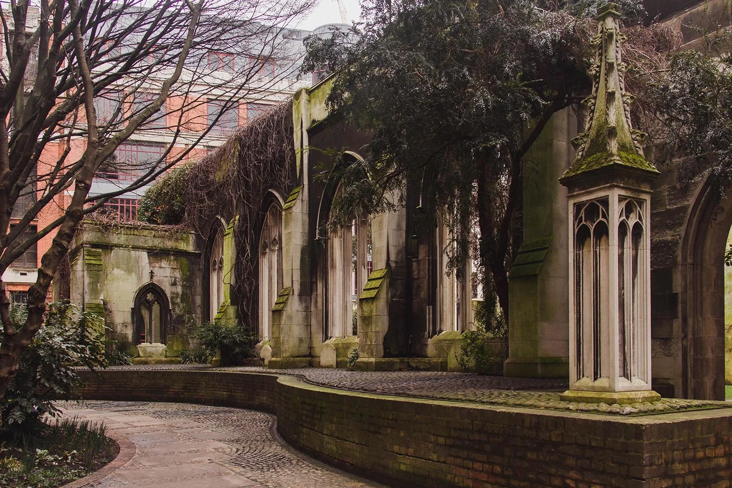 Old stone building with gothic-style arched windows, some covered by overgrown vines, and decorative stone pillars, surrounded by a curved brick pathway and trees, in an urban setting.