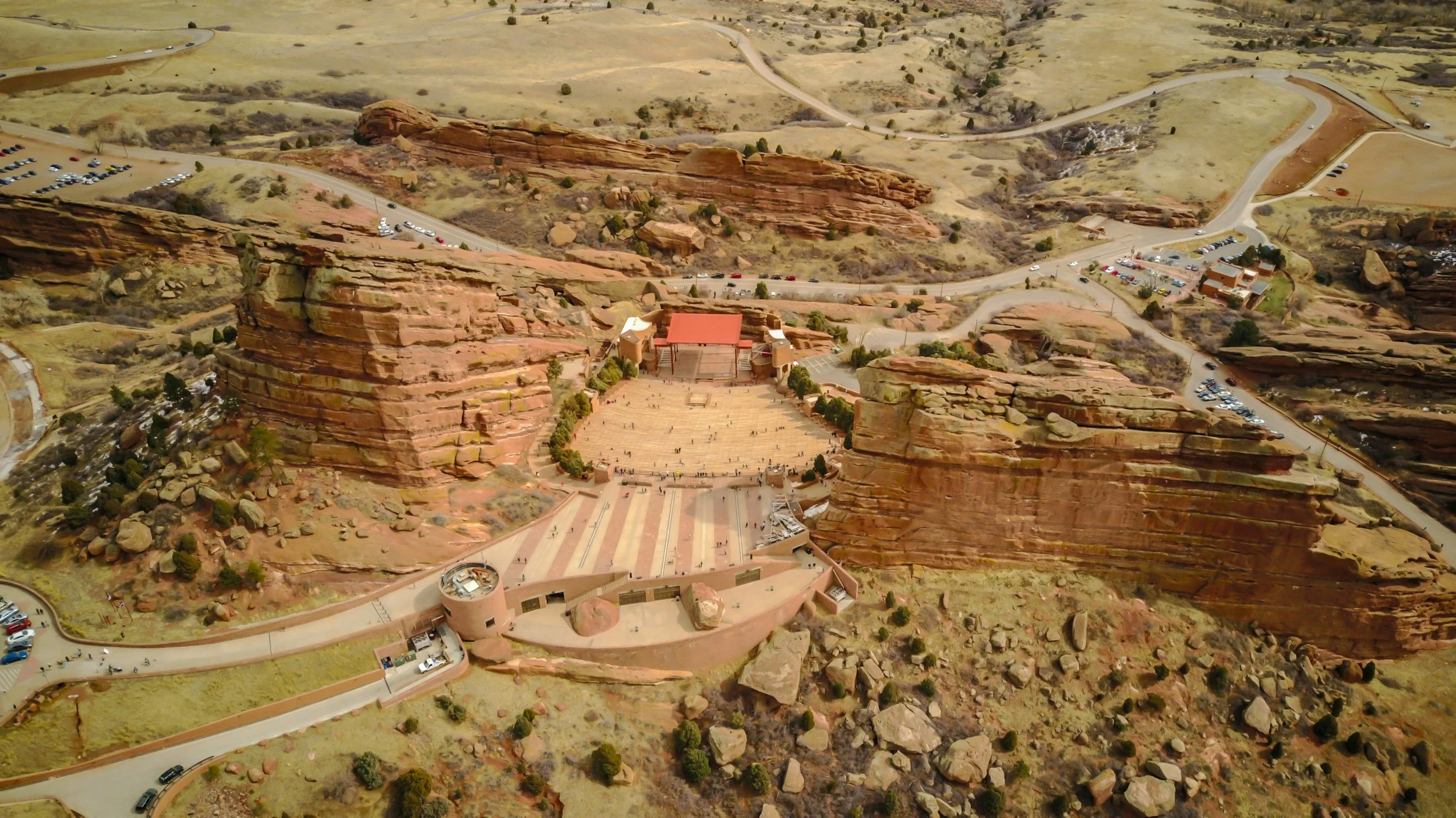Aerial view of Red Rocks Amphitheatre, an outdoor concert venue built into red sandstone rock formations in Colorado, with surrounding parking lots and landscape.