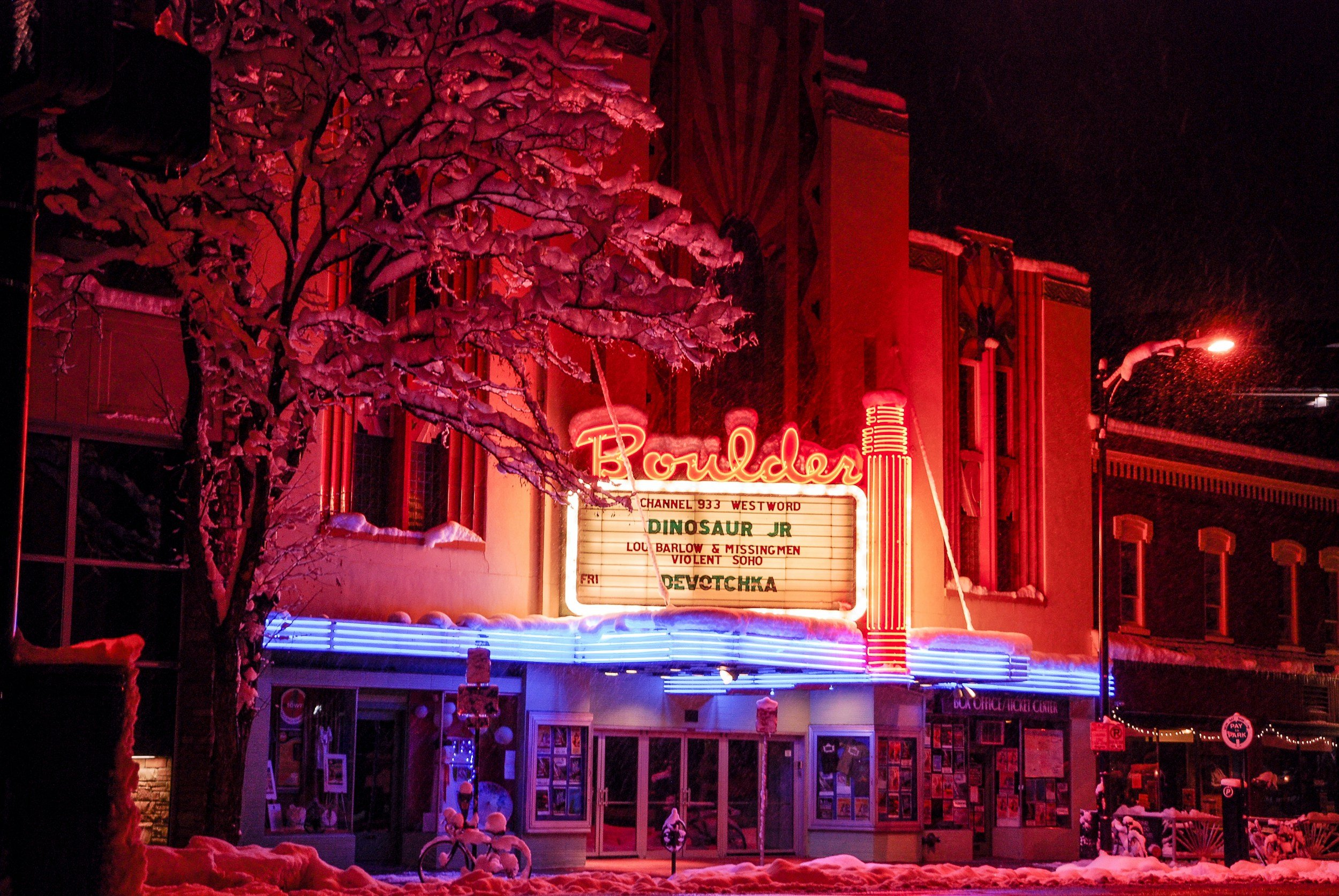 Nighttime scene outside a movie theater named Boulder, with a snow-covered tree in front and neon signs, including a marquee with show listings and a lit sign reading 'Boulder' in red.