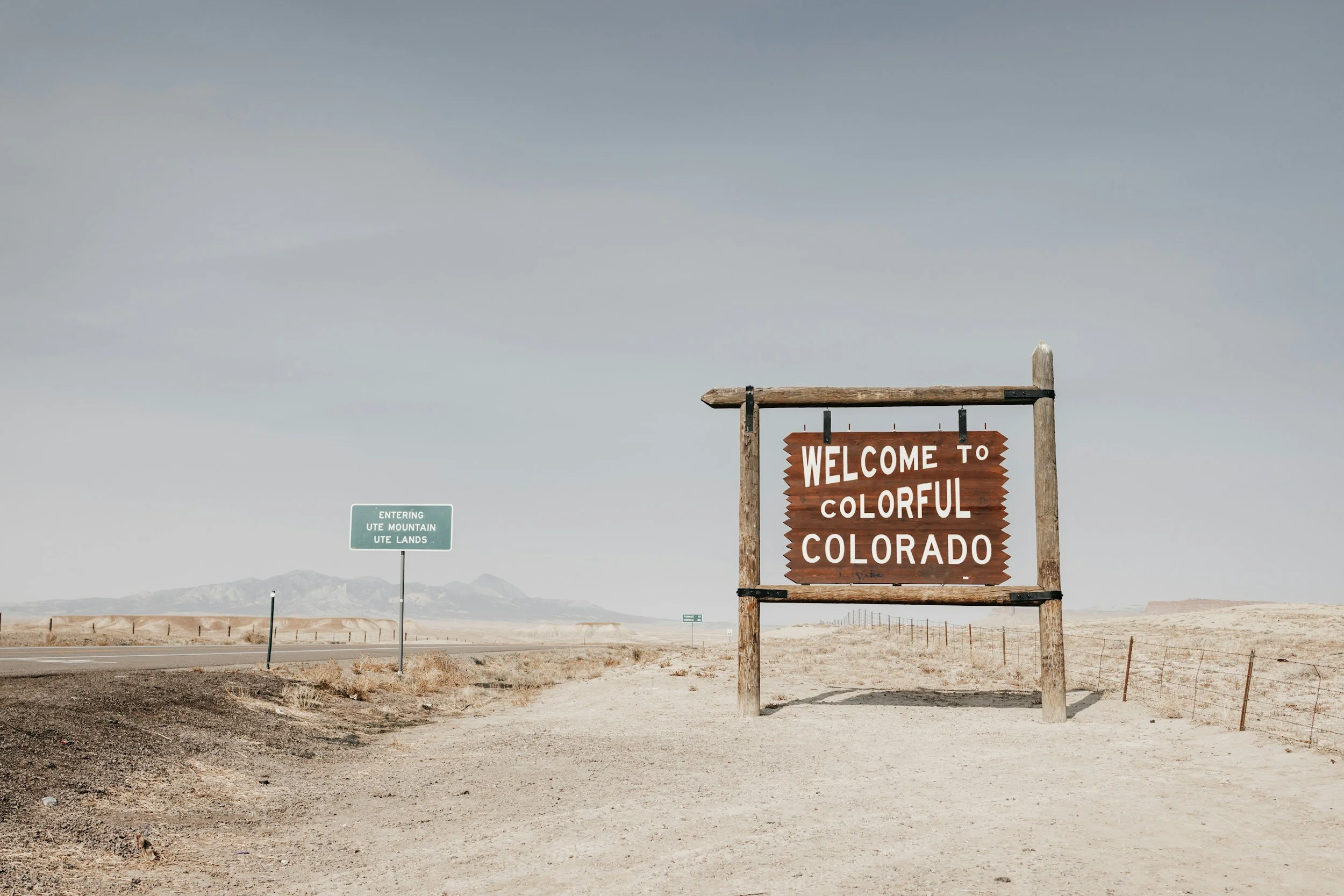 A wooden sign reading 'Welcome to Colorful Colorado' on a dirt roadside in a desert landscape with mountains in the distance and a smaller green sign indicating entry into Ute Mountain Ute Lands.