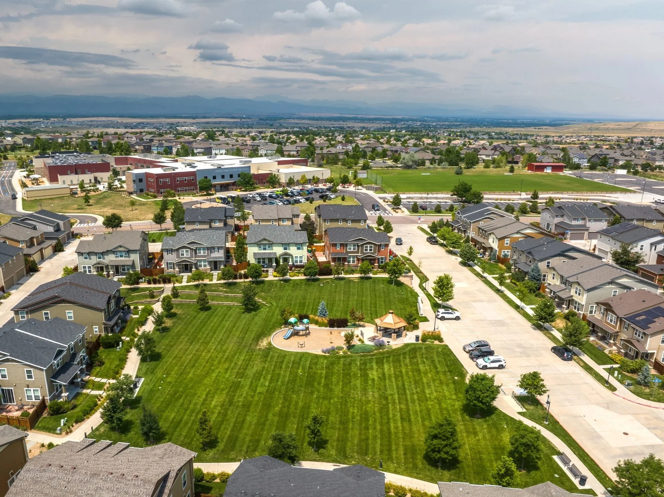 Aerial view of a residential neighborhood with a park, playground, parking lot, and surrounding houses, with mountains and cloudy sky in the background.