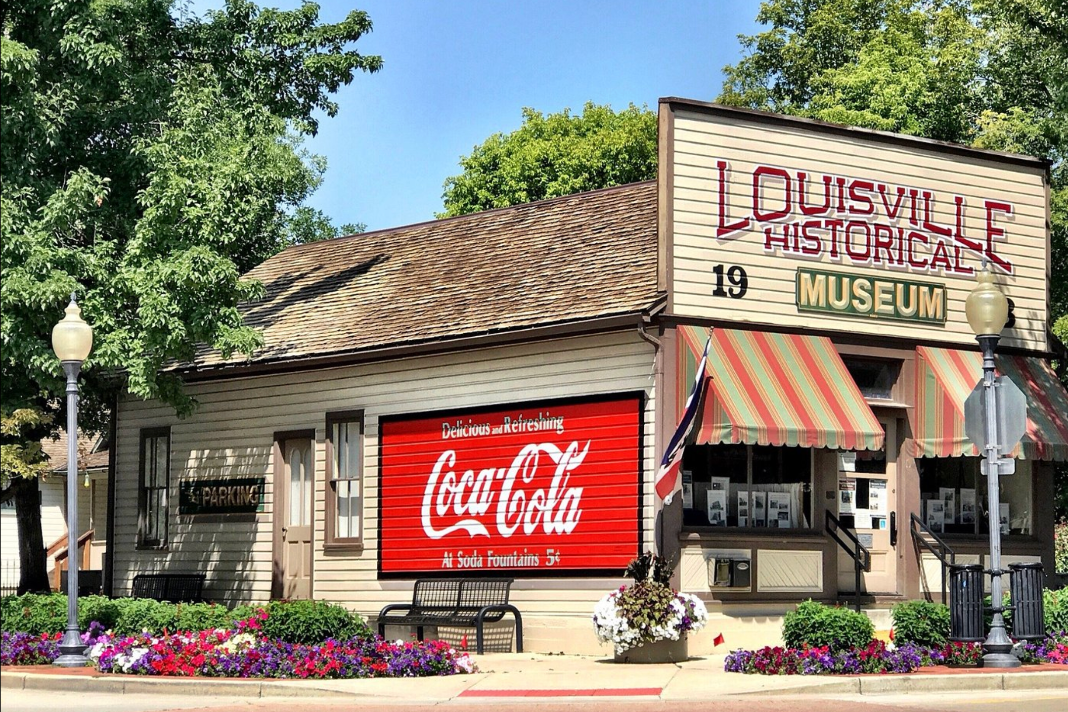 A historic building housing the Louisville Historical Museum with a large sign reading 'Louisville Historiale Museum' and the year 1919. The building has striped awnings over the entrance, flower beds with colorful flowers, a Coca-Cola sign, and street lamps, with trees and a clear blue sky in the background.