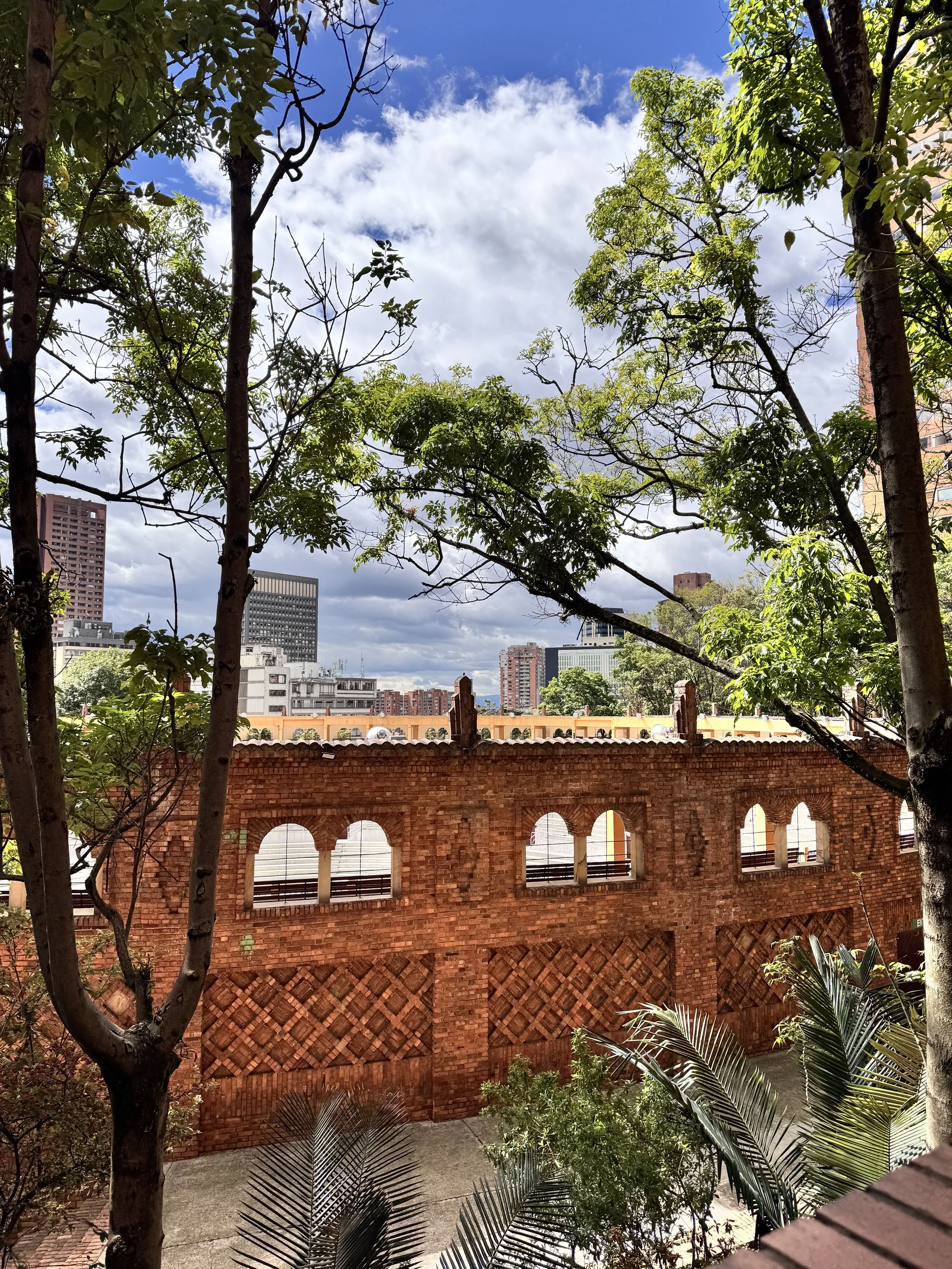Cityscape with trees and a red brick wall in the foreground, and modern buildings against a partly cloudy sky in the background.