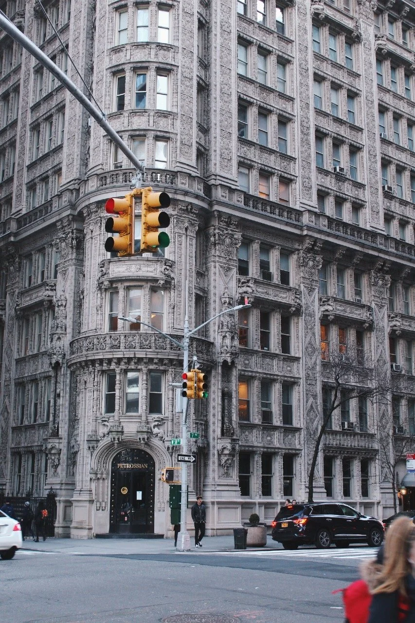 A city street intersection with traffic lights, a historic multi-story building with ornate architecture, and pedestrians walking nearby.