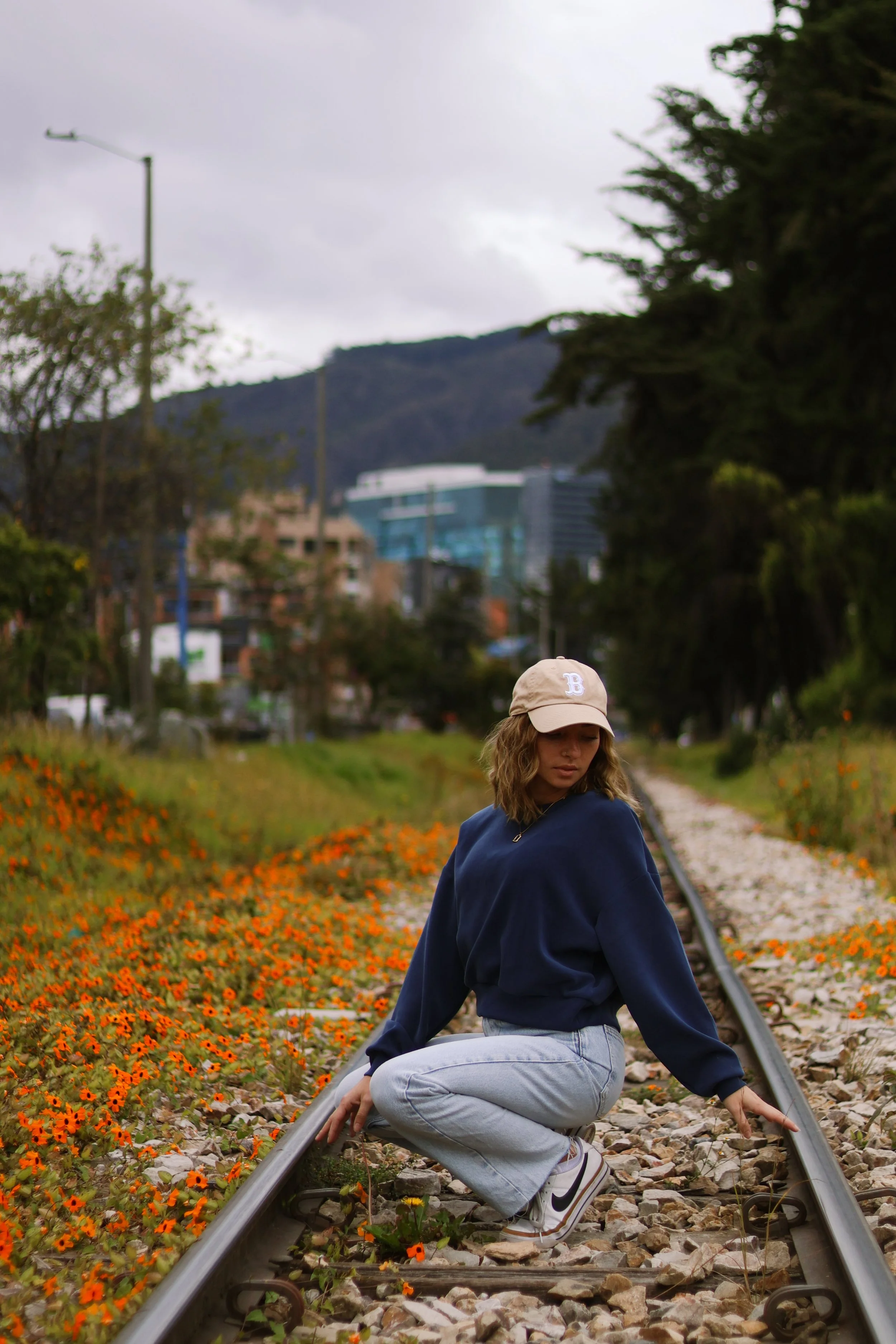 A woman crouching on train tracks surrounded by orange flowers with city buildings and hills in the background during overcast weather.