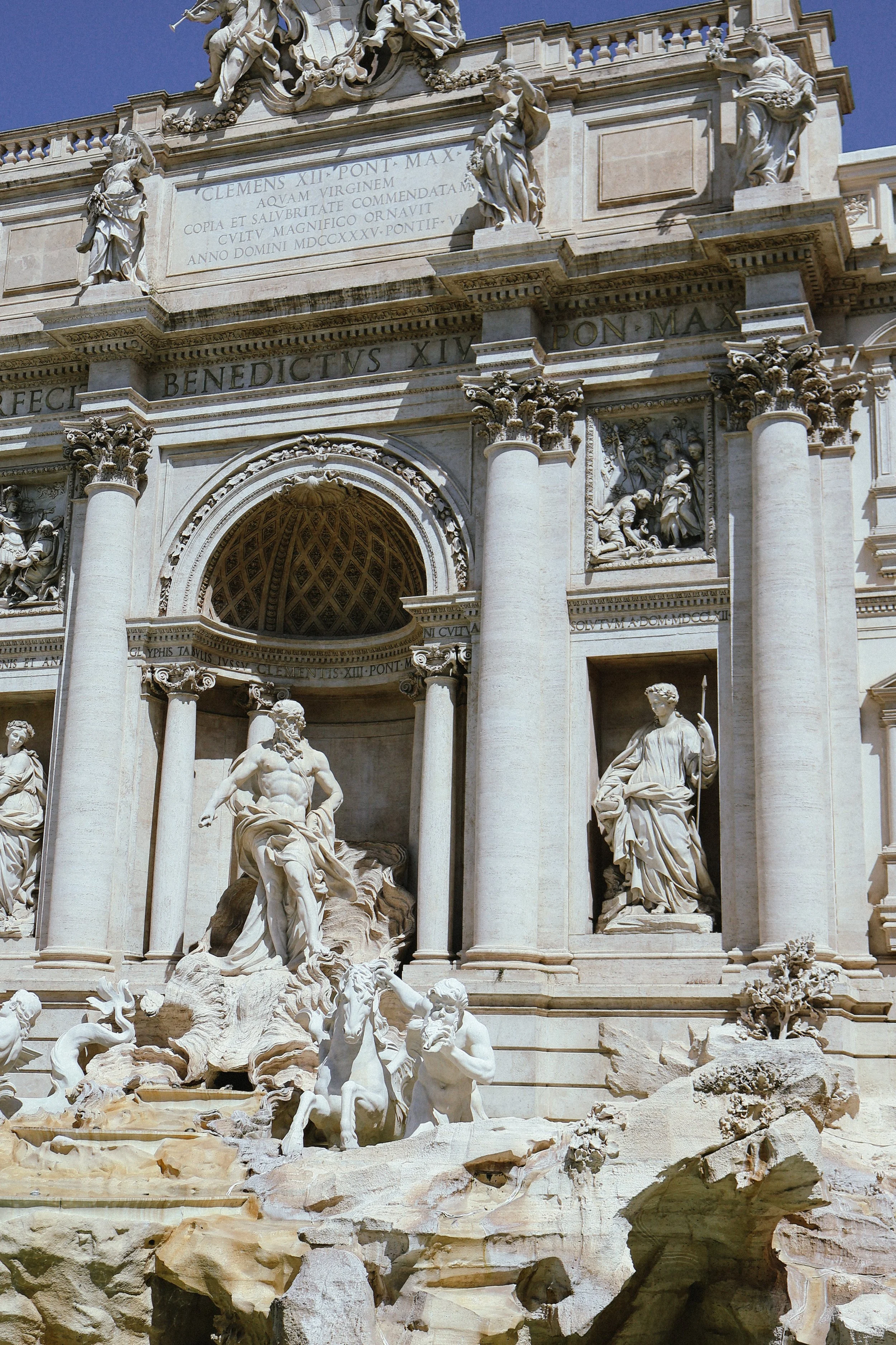 The Trevi Fountain in Rome, Italy, featuring elaborate sculptures of mythological figures, including a central figure of a man with a beard, surrounded by multiple columns, statues of figures, and rocky landscapes with water features.