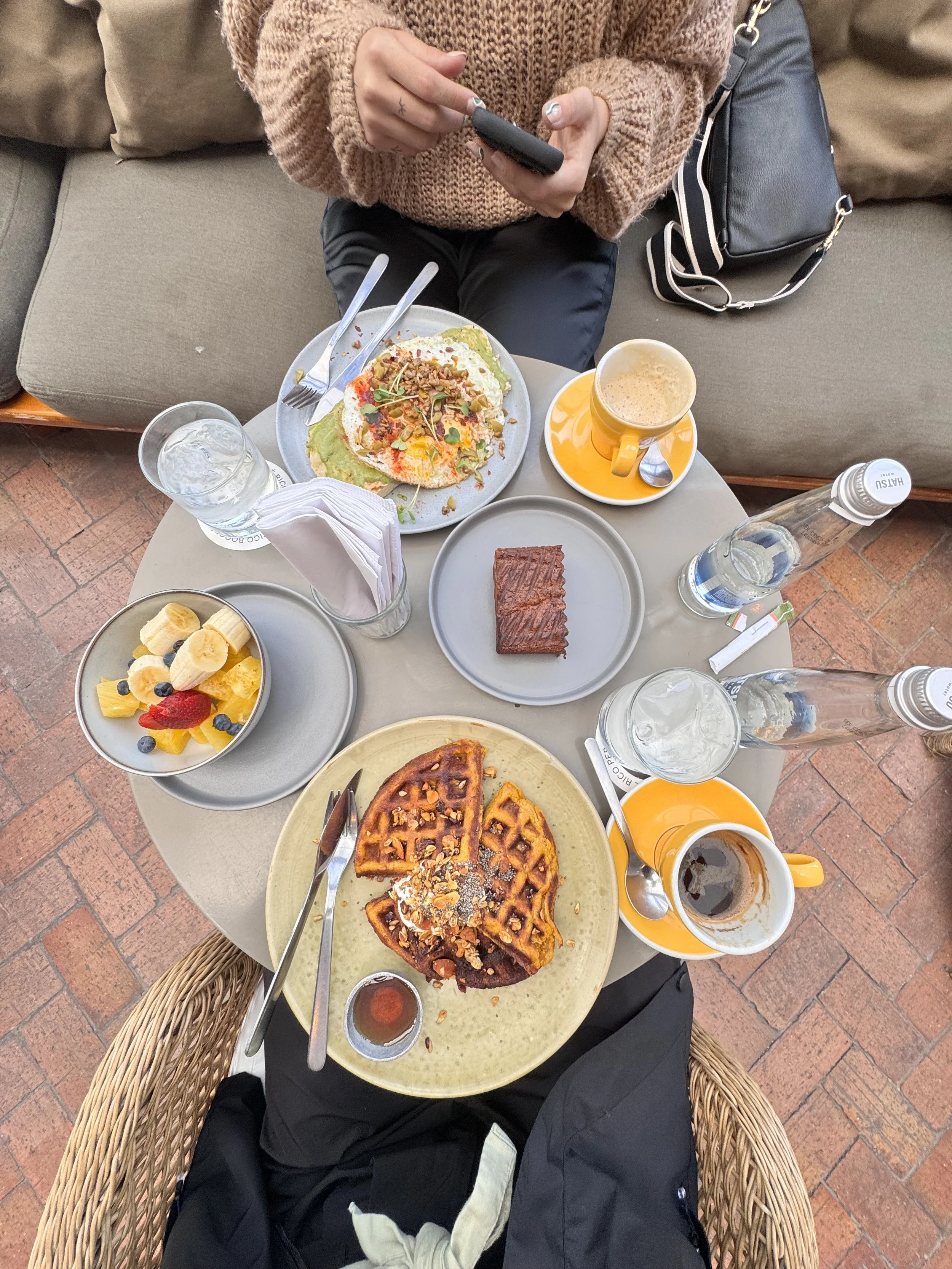 Top-down view of a table with waffles, fruit salad, chocolate baked good, coffee, water bottles, and a person using a phone.