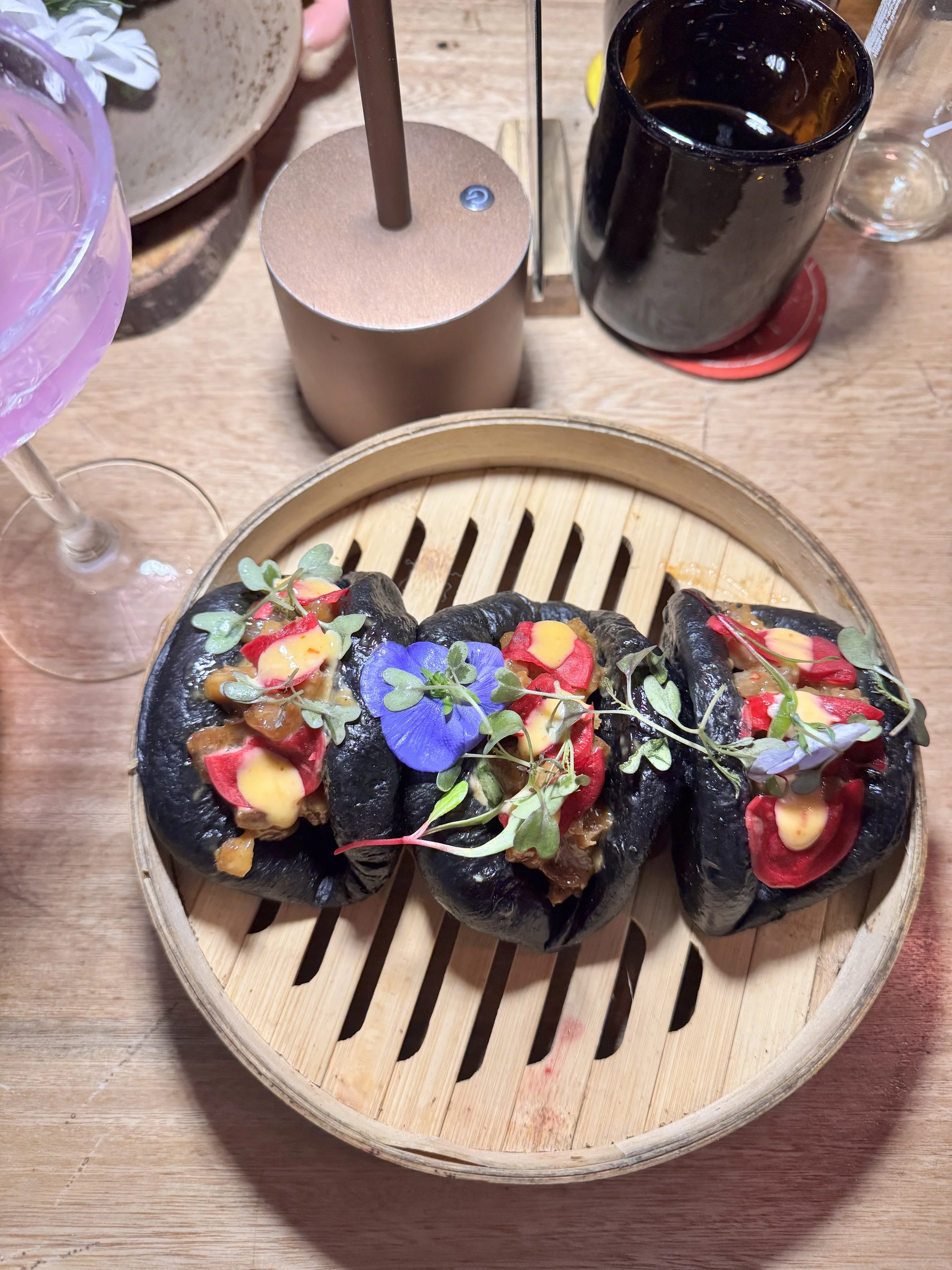 Three black steam buns filled with meat and topped with microgreens, edible flowers, and colorful sauces, served on a round bamboo steamer.