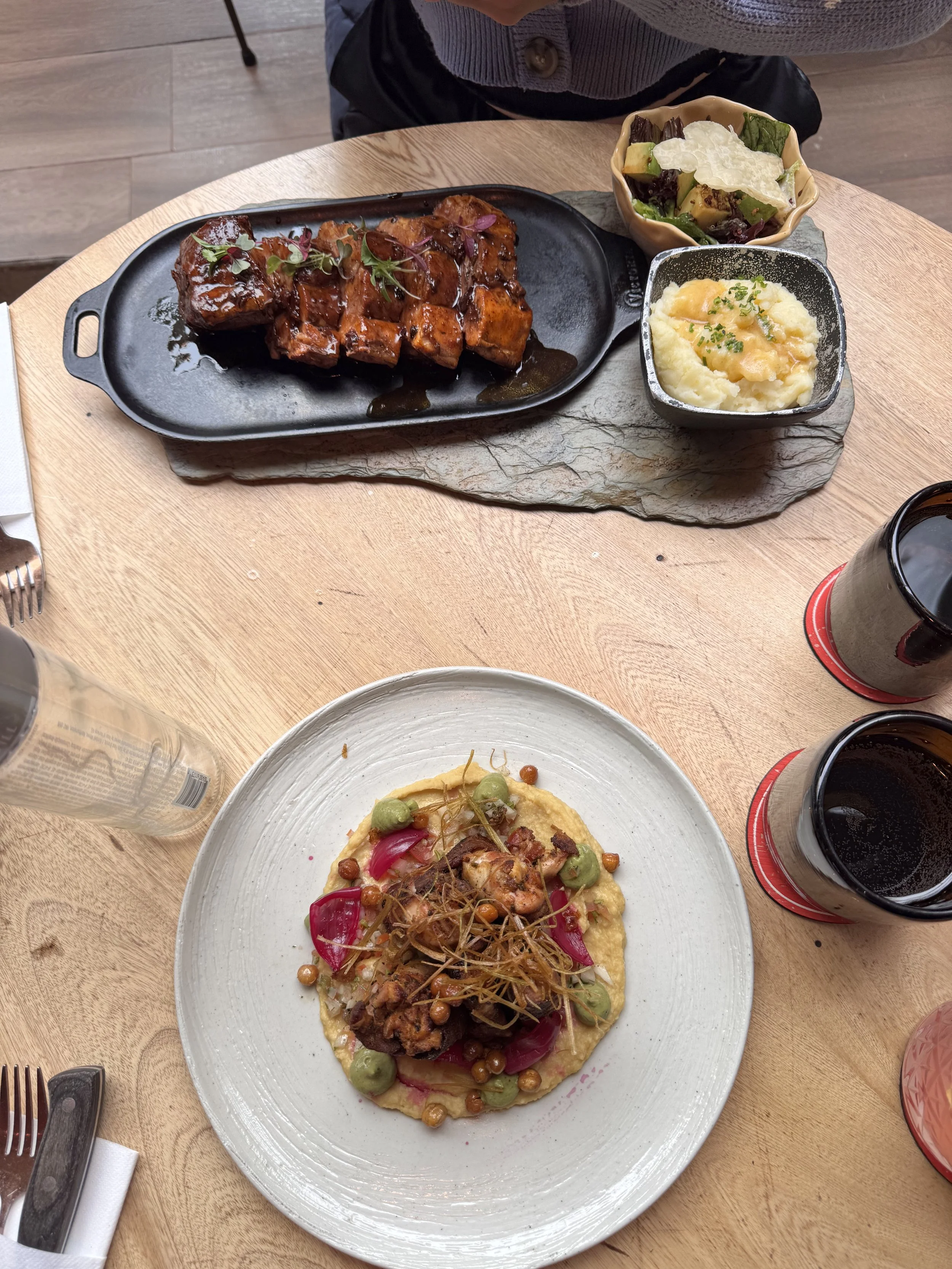 A meal on a wooden table with two plates of food, a small bowl of salad, a small bowl of mashed potatoes, and glasses of dark soda.