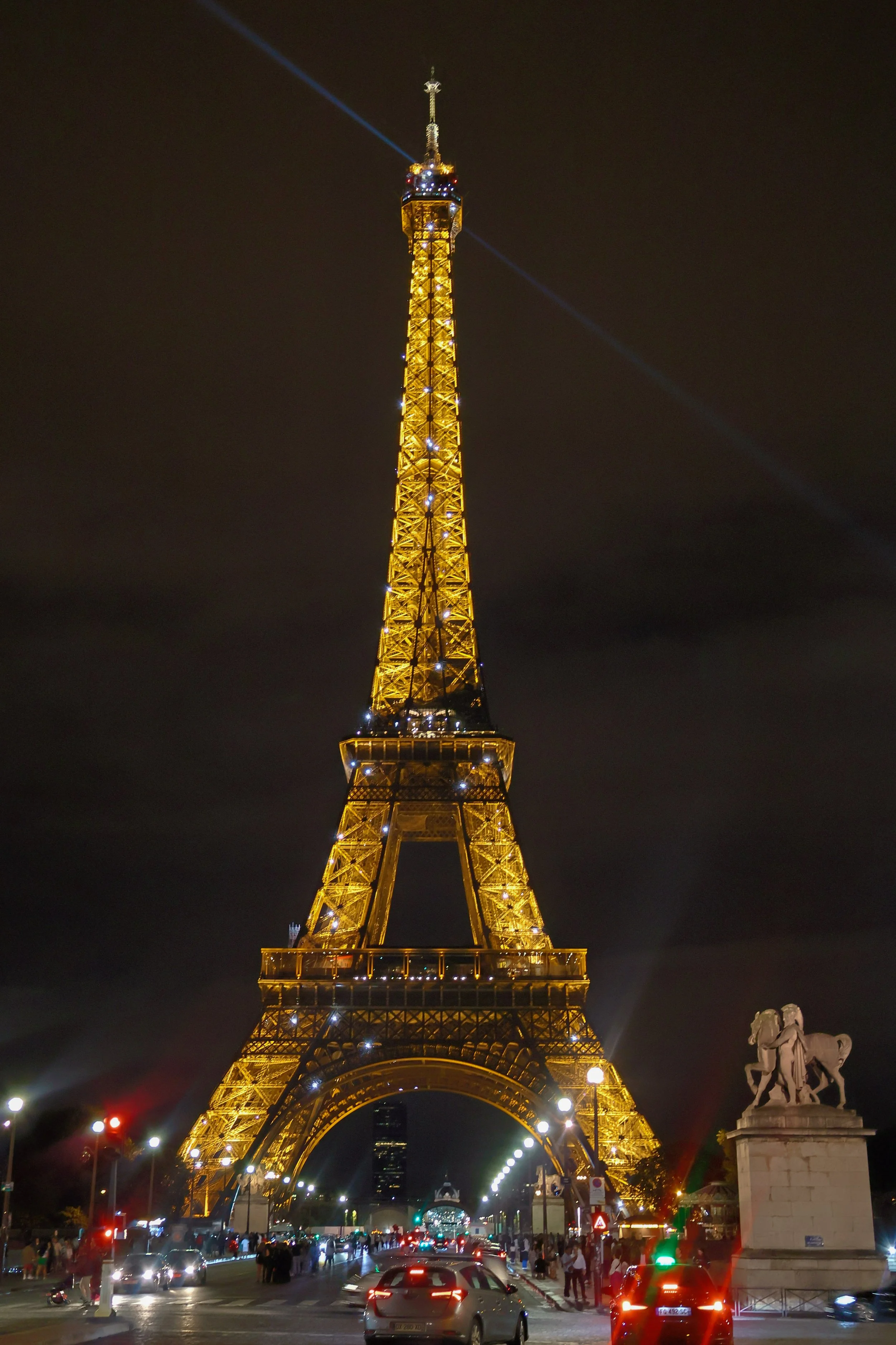Night view of the Eiffel Tower in Paris illuminated with golden lights, with cars and people below and a statue on the right.