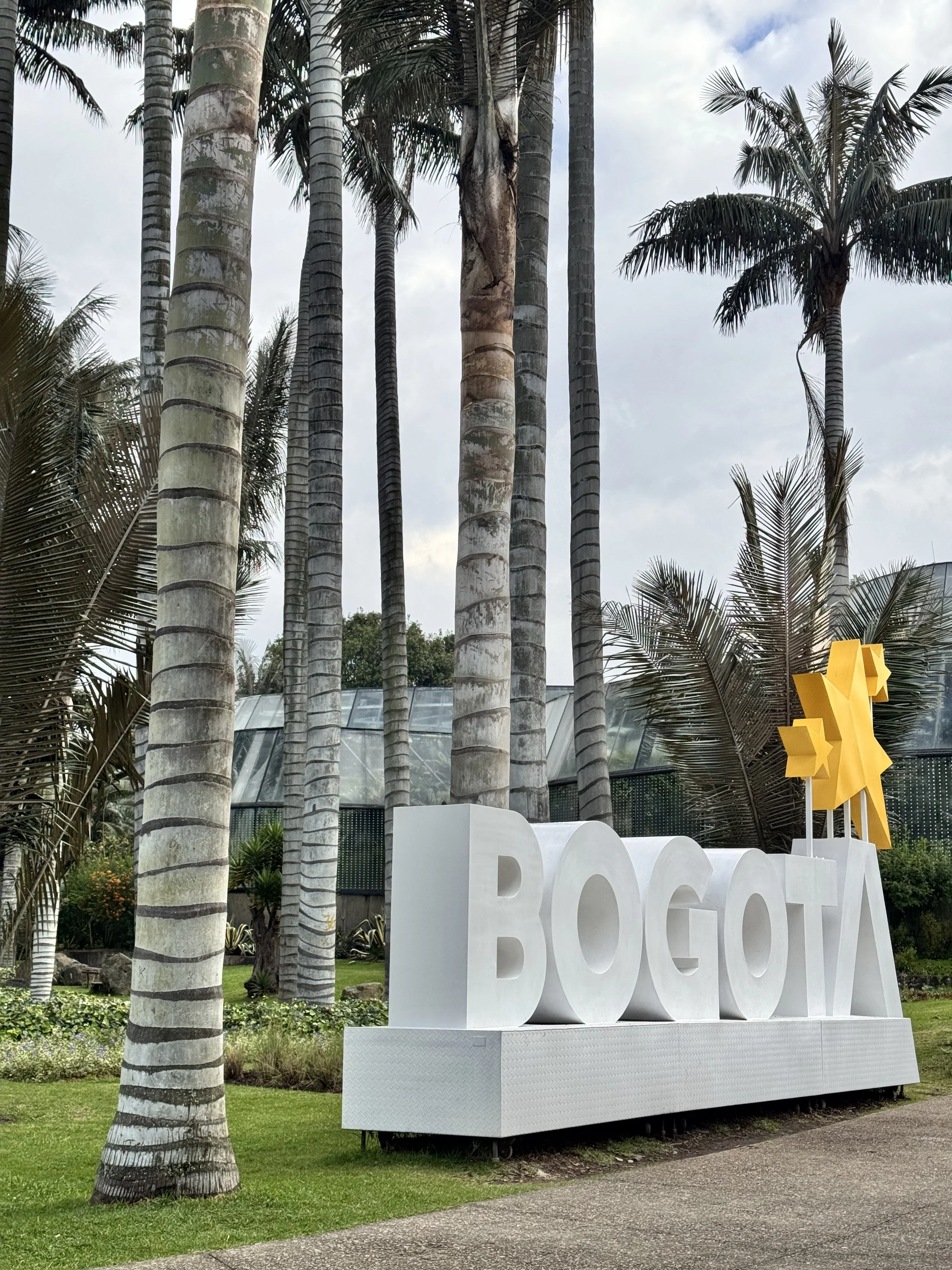 Large white sign spelling out 'BOGOTÁ' with a yellow abstract sculpture on top, surrounded by tall palm trees and lush greenery in an outdoor park setting.