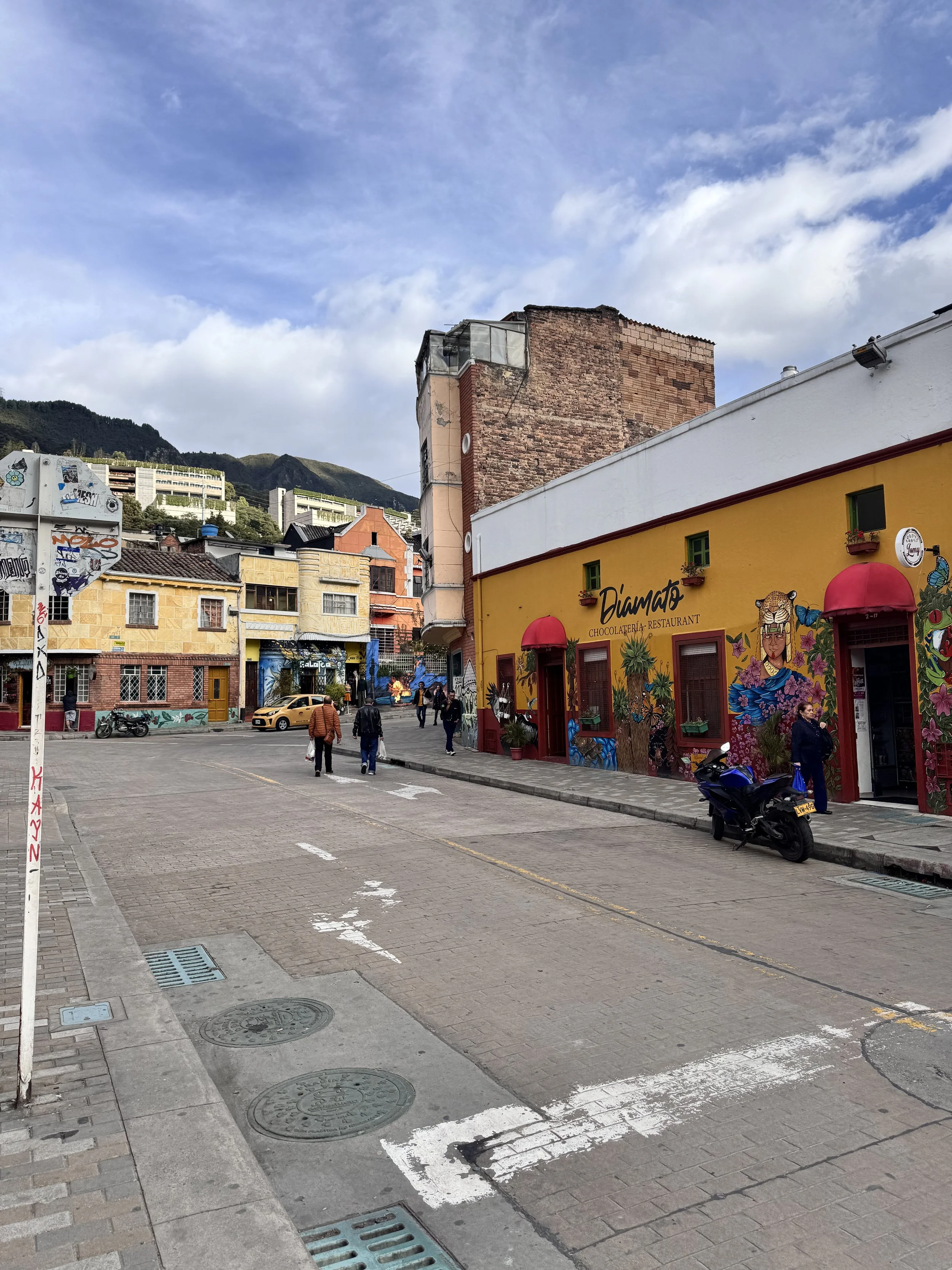 Colorful street scene with pedestrians, motorcycles, and vibrant murals on buildings, including a yellow restaurant called "Diamato" with red awnings and decorative artwork, under a partly cloudy sky with mountains in the background.